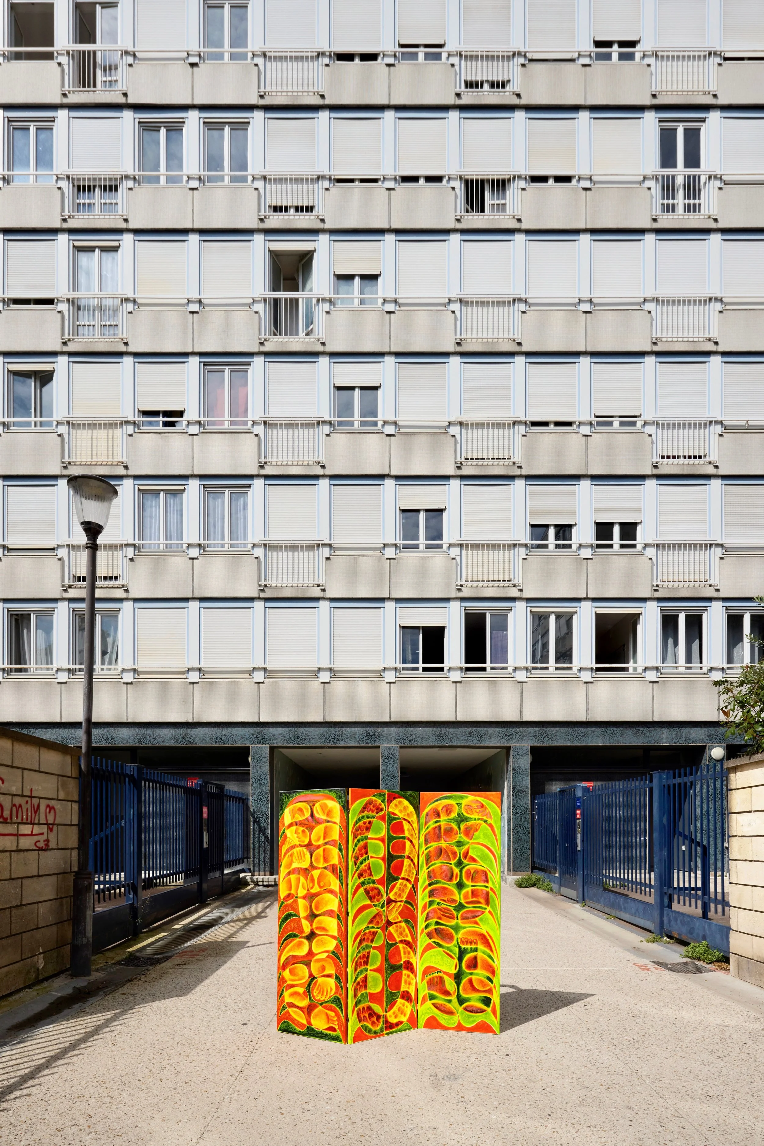 Colorful abstract art sculpture with bright yellow, orange, and green patterns at the entrance of a modern residential building with closed windows and balconies.