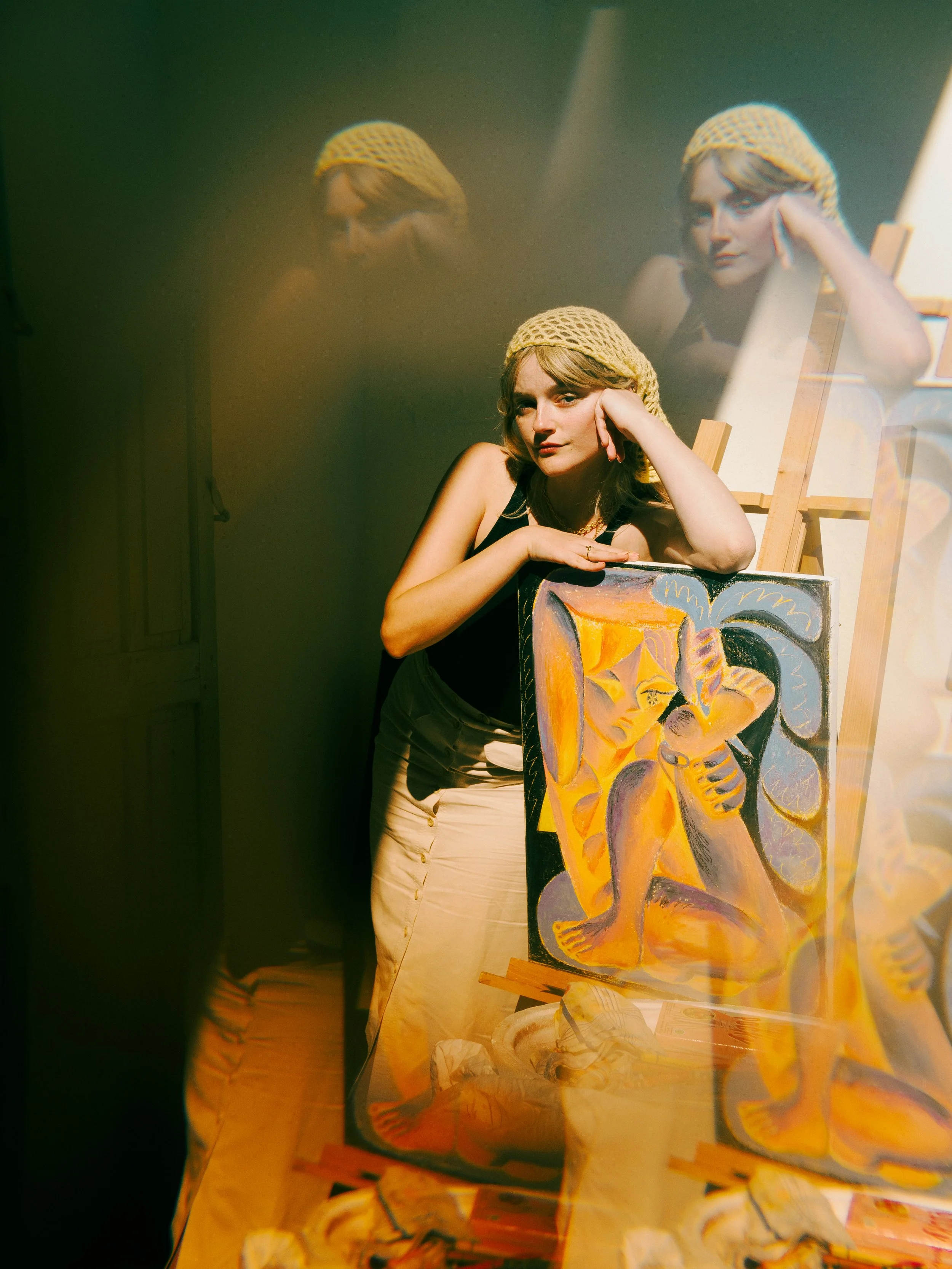A woman wearing a yellow knit hat resting her face on her hand, with a colorful abstract painting in the foreground and a mirror reflecting her face behind her.