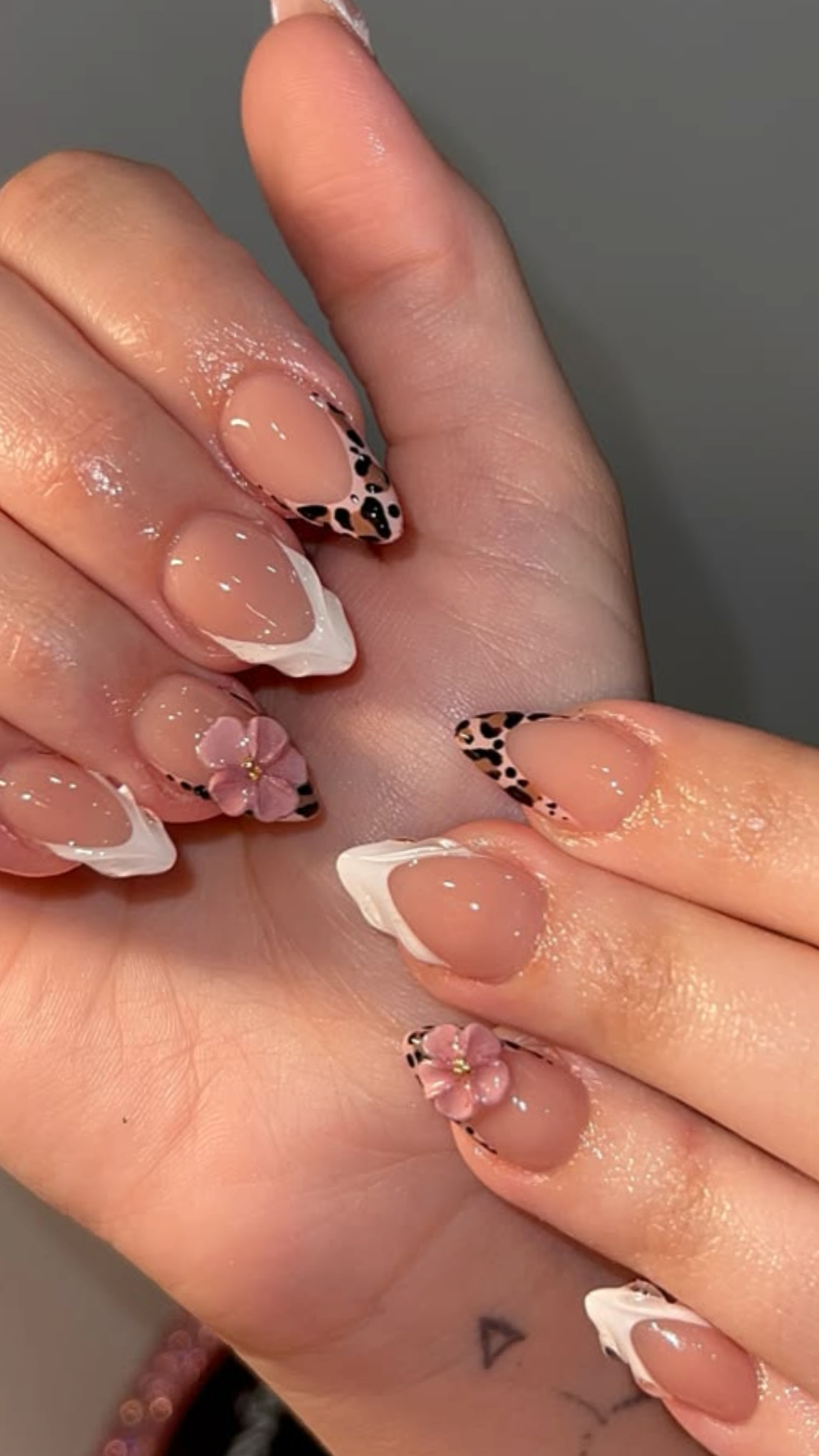 Close-up of manicured nails with leopard print and pink flower designs, held in a person's hand with a small triangle tattoo visible.