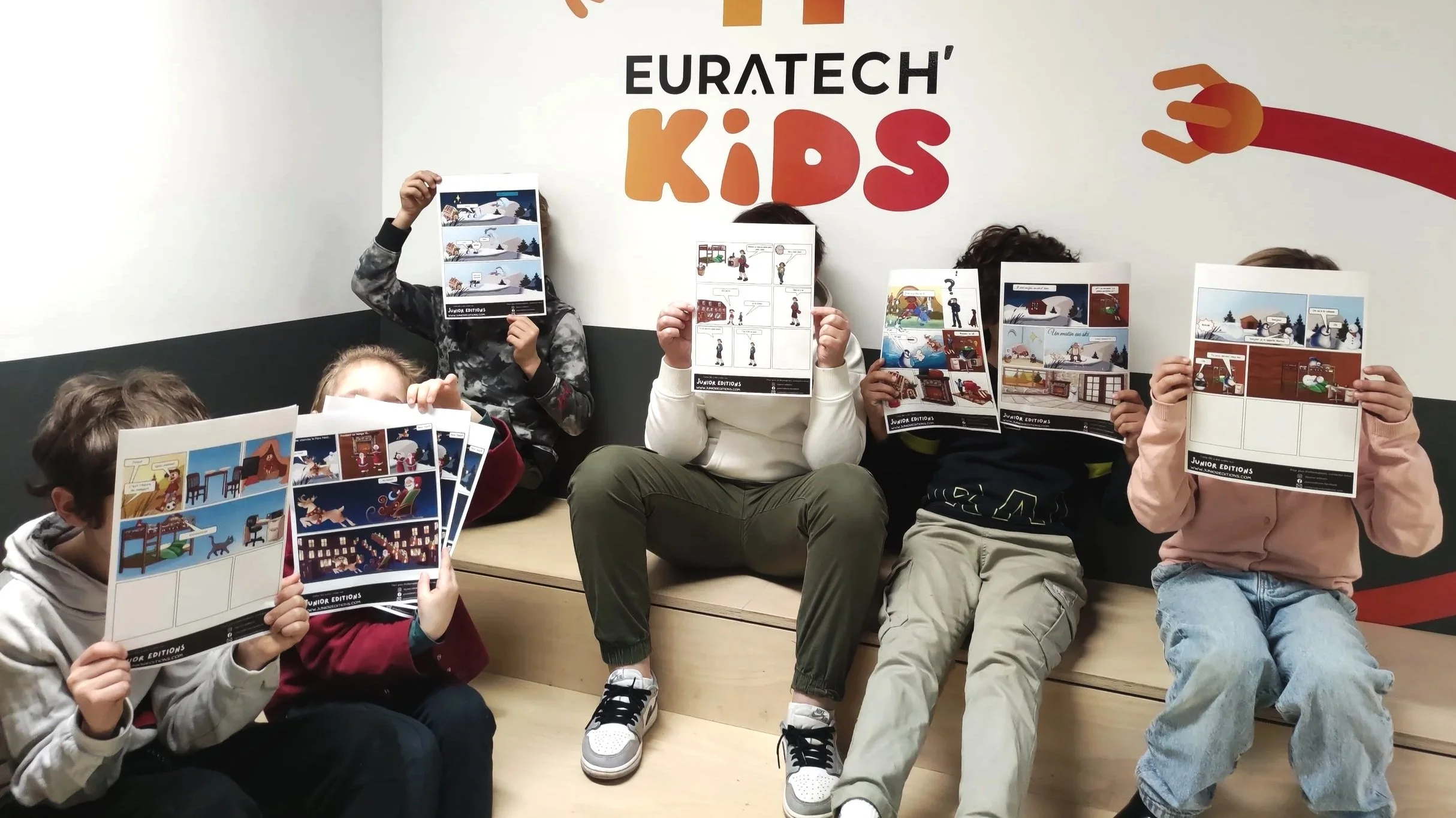 Groupe d'enfants assis contre un mur blanc portant une affiche 'EURATECH KIDS' en arrière-plan, chacun tenant une fiche imprimée de bande dessinée avec des scènes diverses.