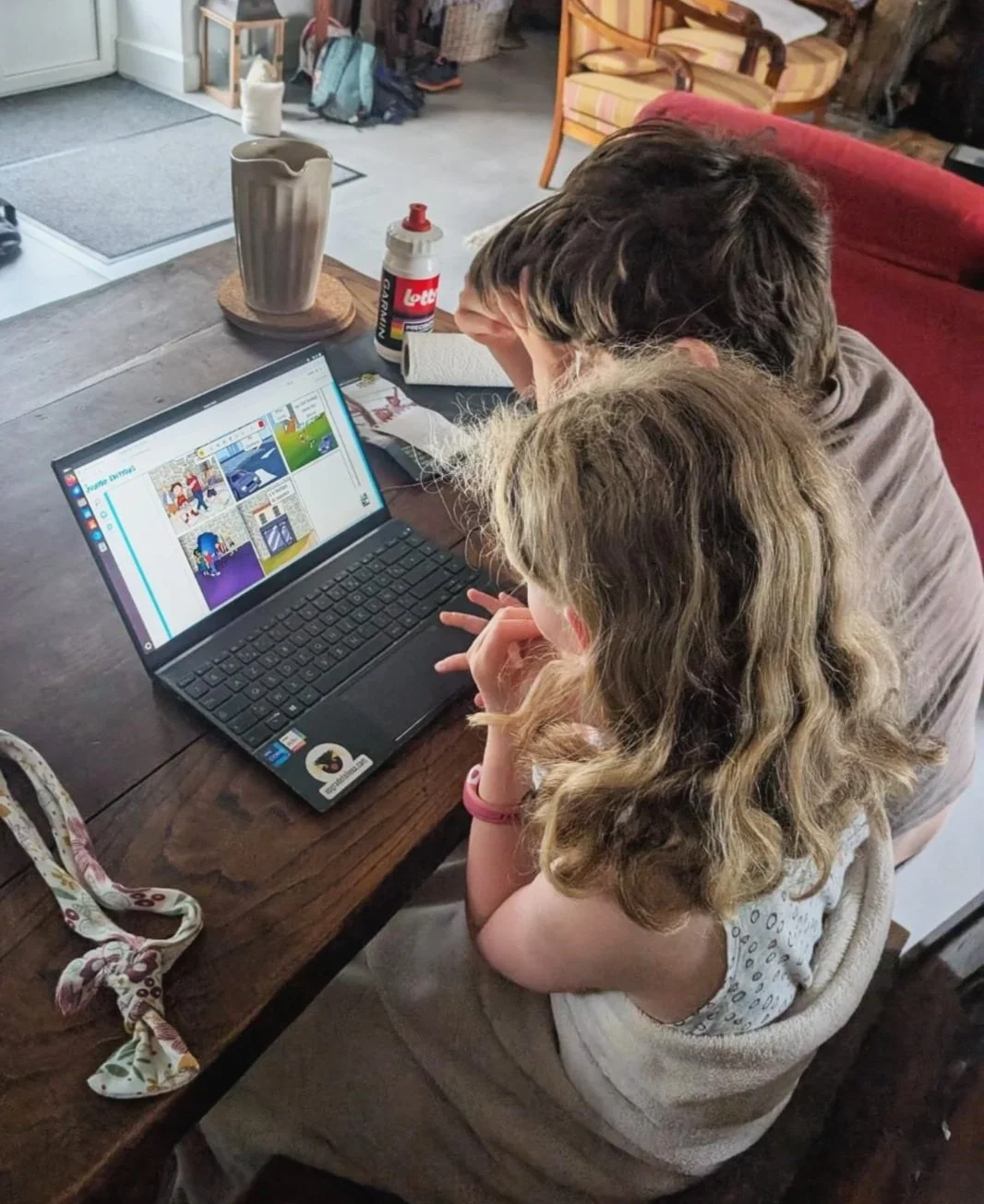 Deux enfants regardent un ordinateur portable sur une table en bois, dans une pièce lumineuse avec des meubles et des objets divers. La scène semble se dérouler à la maison, avec un décor confortable.