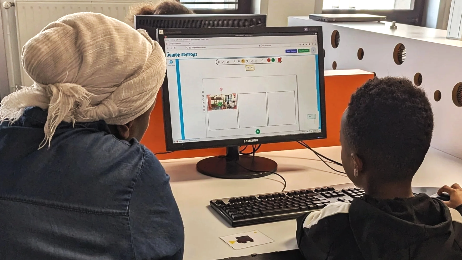 Une maman et son fils travaillent ensemble sur un ordinateur à un bureau, en train de créer une mise en page pour un livre ou une publication, dans une salle informatique de la médiathèque.