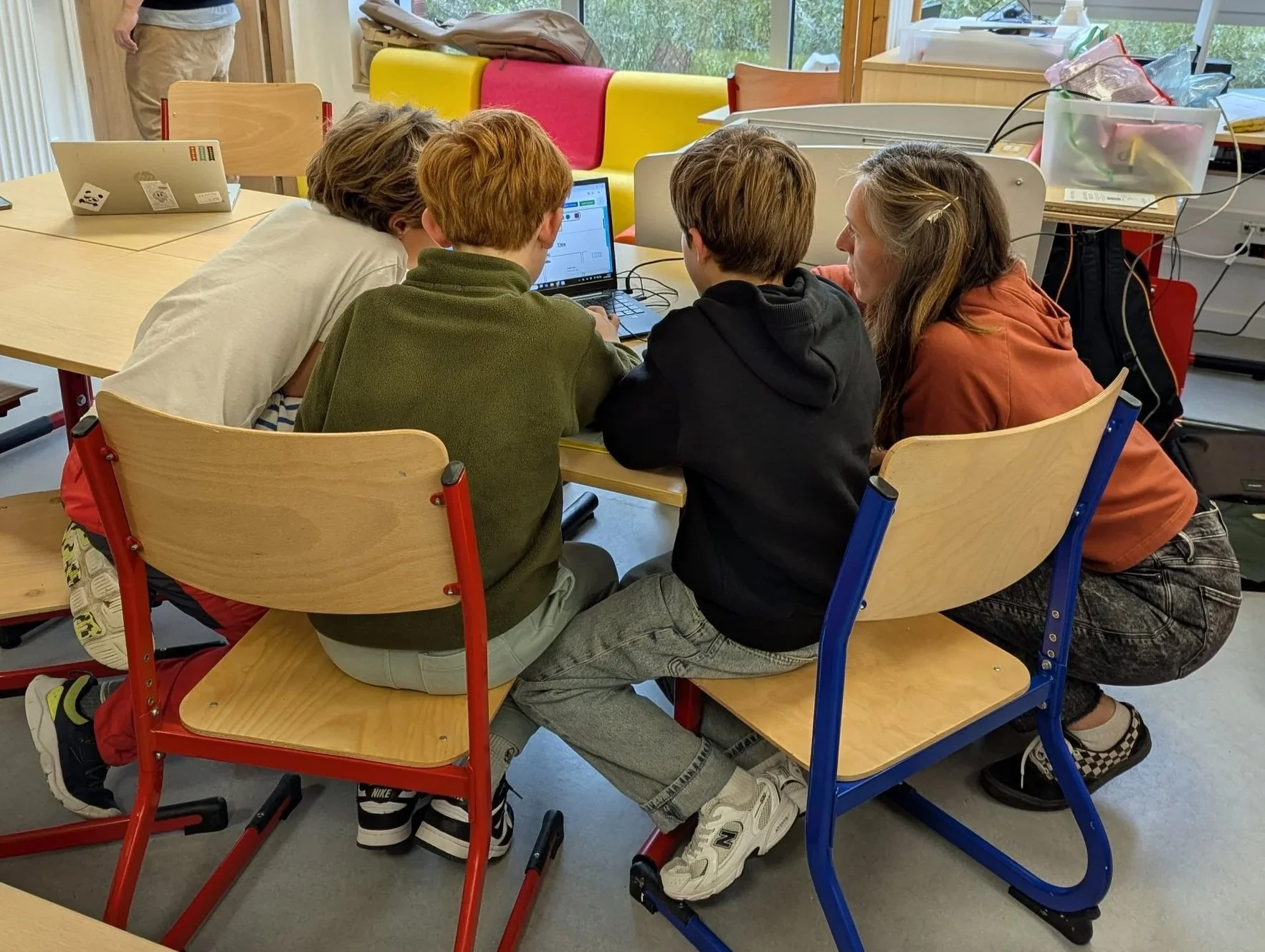 Cinq jeunes assis sur une table regardant un ordinateur portable ensemble avec leur enseignante. La salle est lumineuse avec de grandes fenêtres arrière et des chaises colorées.