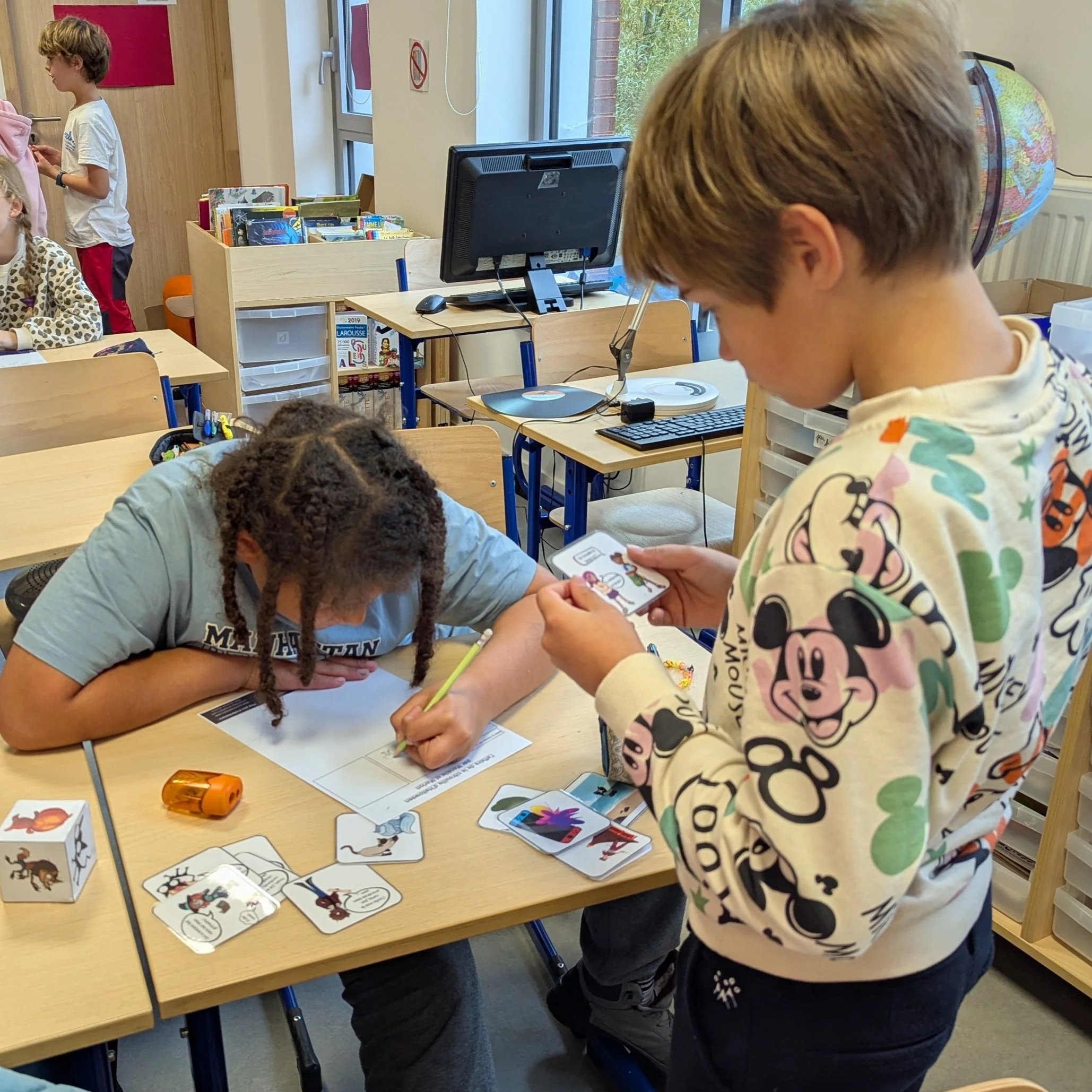 Une classe avec des enfants qui jouent avec des cartes illustrées de différentes créatures et personnages dit lanceurs d'écriture. Certains enfants sont assis à une table ou debout, concentrés ou interagissant.