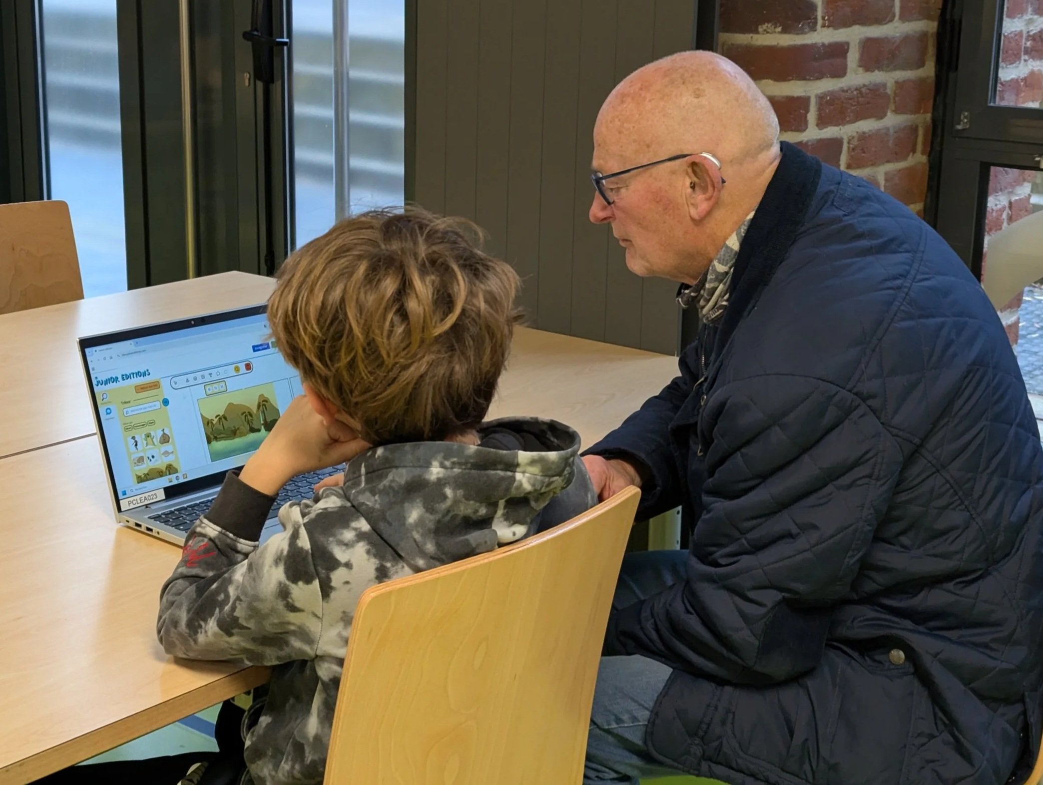 Un grand-père et son petit garçon assis à une table, regardant un ordinateur portable en intérieur, en plein création de BD numérique