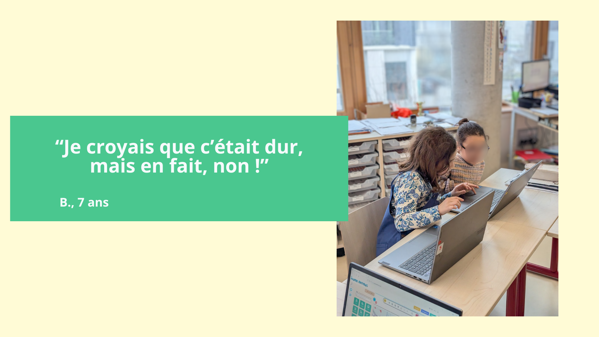 Deux enfants assis à une table d'école travaillent sur des ordinateurs portables dans une classe. La pièce dispose de grandes fenêtres et de rangements avec des tiroirs. Un des enfants porte un sweat à manches longues à motifs bleus.