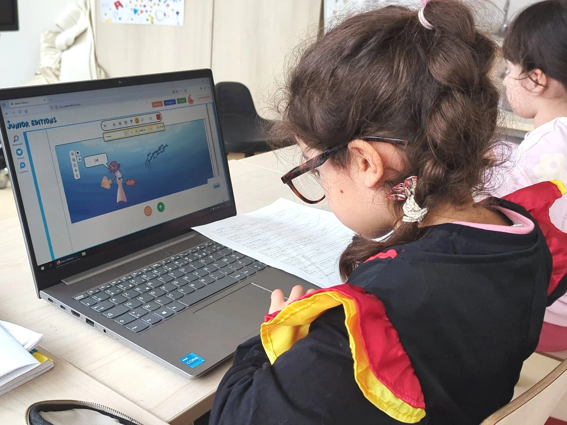 Une jeune fille portant un ourlet rouge, jaune et noir travaille devant un ordinateur portable dans une salle de classe. L'écran de l'ordinateur affiche un site web éducatif pour enfants.