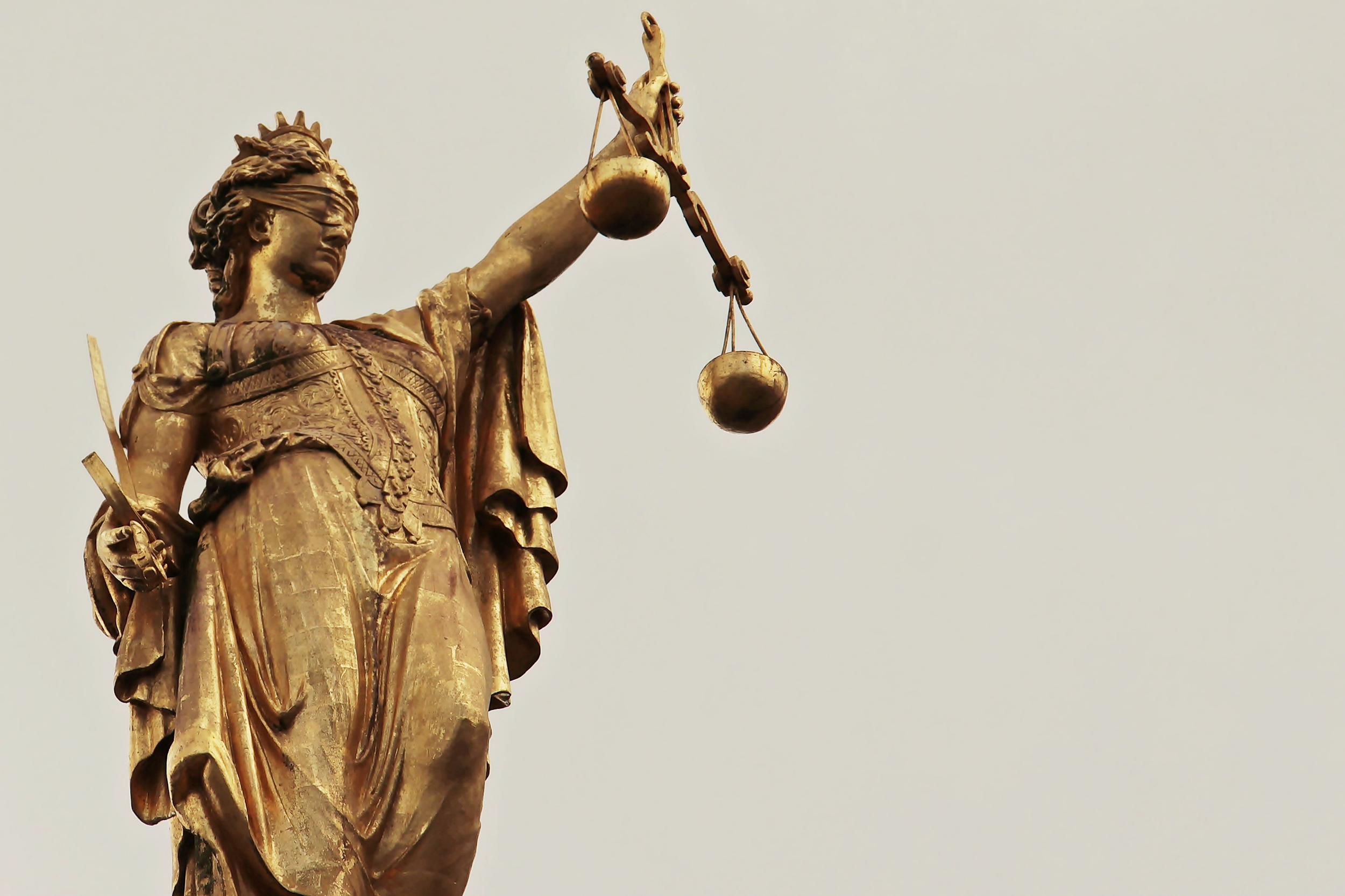 A golden statue of Lady Justice holding a set of scales in her left hand, with her right hand resting at her side, against a plain sky background.