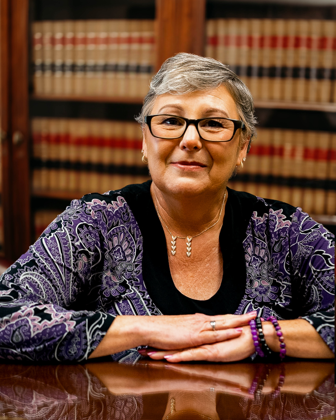 A smiling woman, Stephanie Jones, with short gray hair and glasses, sitting at a polished wooden table in front of bookshelves filled with legal books, wearing a patterned purple and black blouse and jewelry.