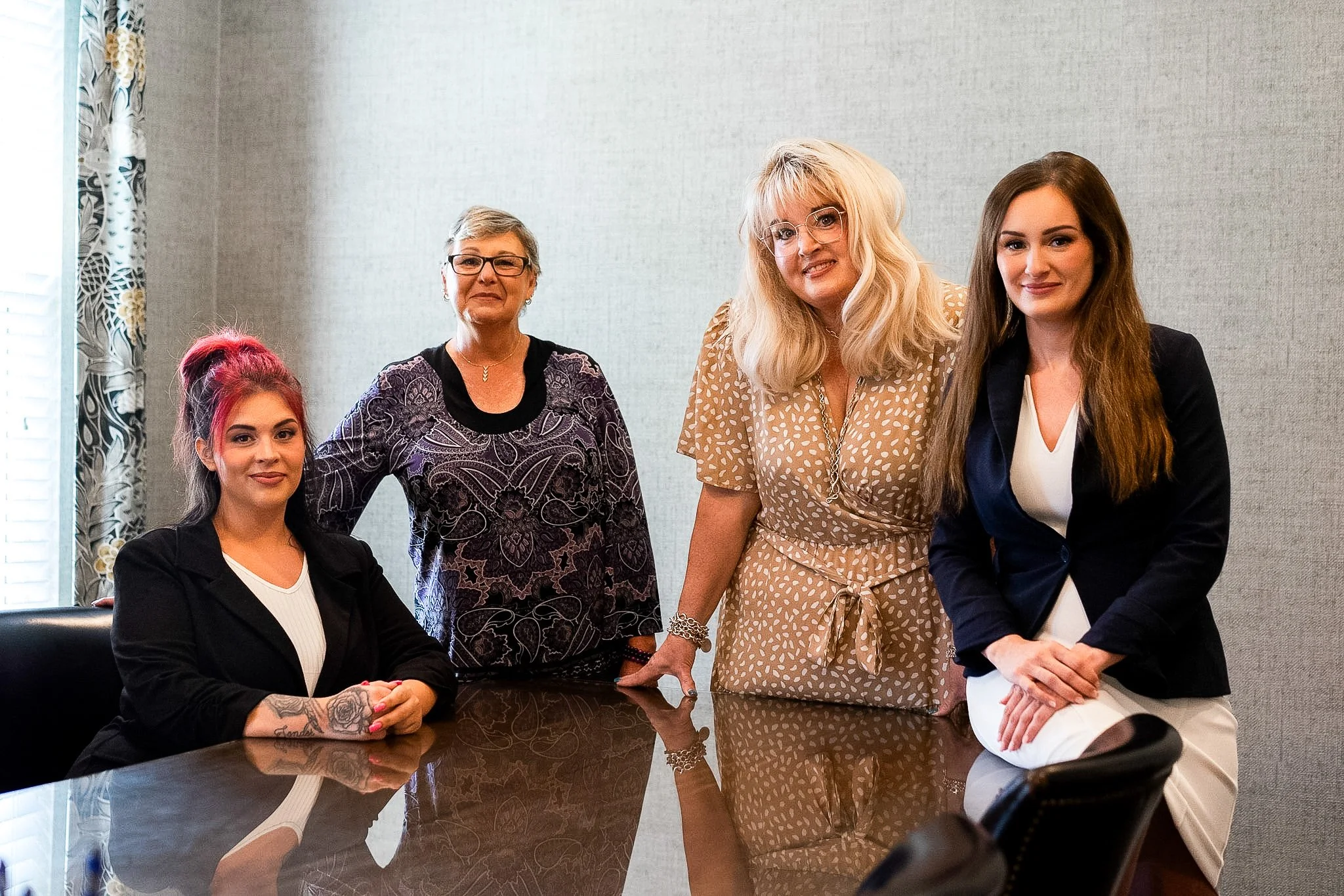 Group of four women in the Peil Law team in a professional setting. Two are seated at a dark wood table, and three are standing behind them. The women vary in age and appearance, dressed in business attire, with smiles and confident expressions.