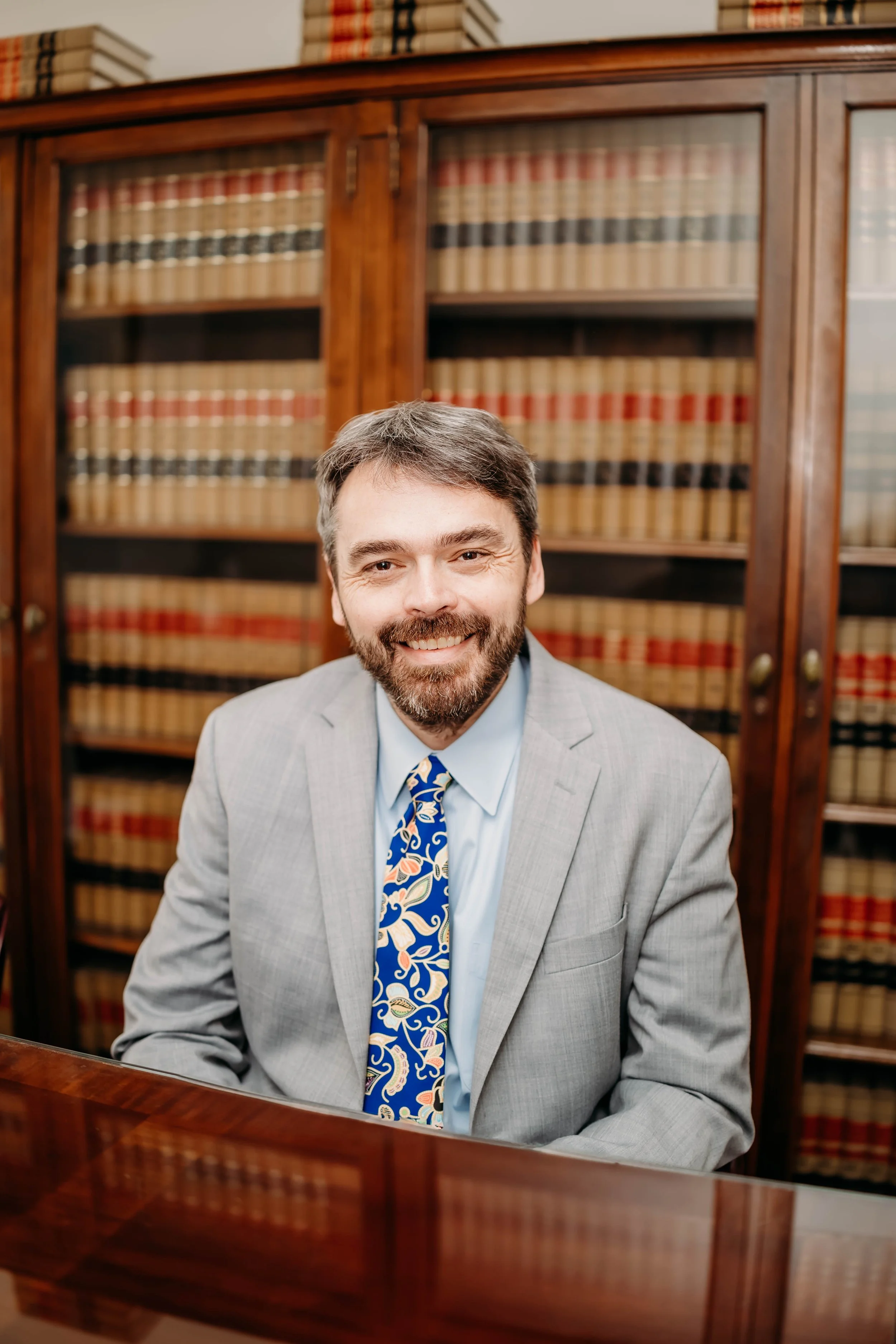 A man, Jeff Peil, with dark hair and a beard smiling, wearing a light gray suit and a blue tie with colorful pattern, seated in front of a wooden bookshelf filled with legal books.