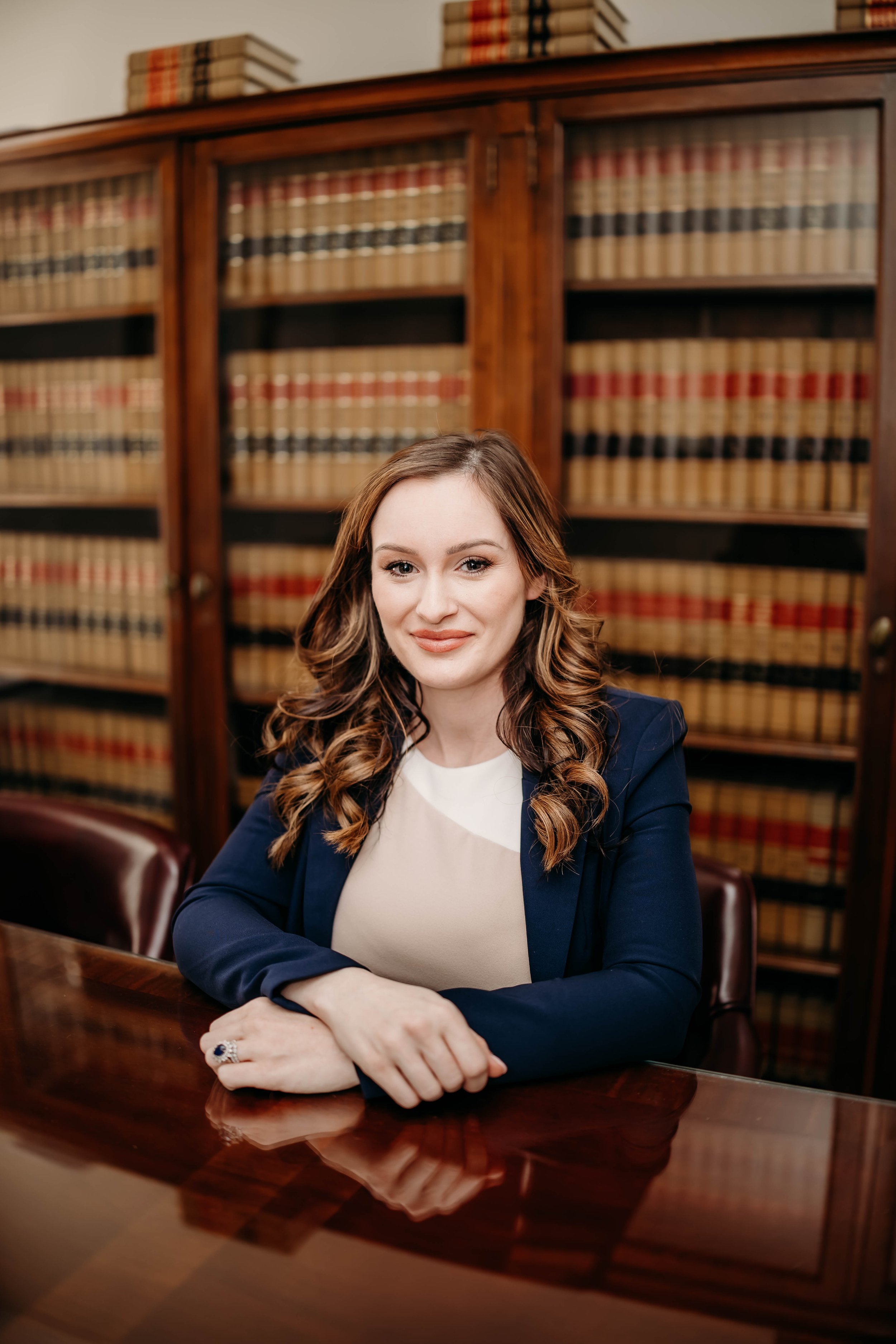A woman, Brianna Peil, with wavy brown hair, wearing a navy blazer and white top, sitting at a polished wooden desk in a library with wooden bookshelves filled with legal or law books in the background.