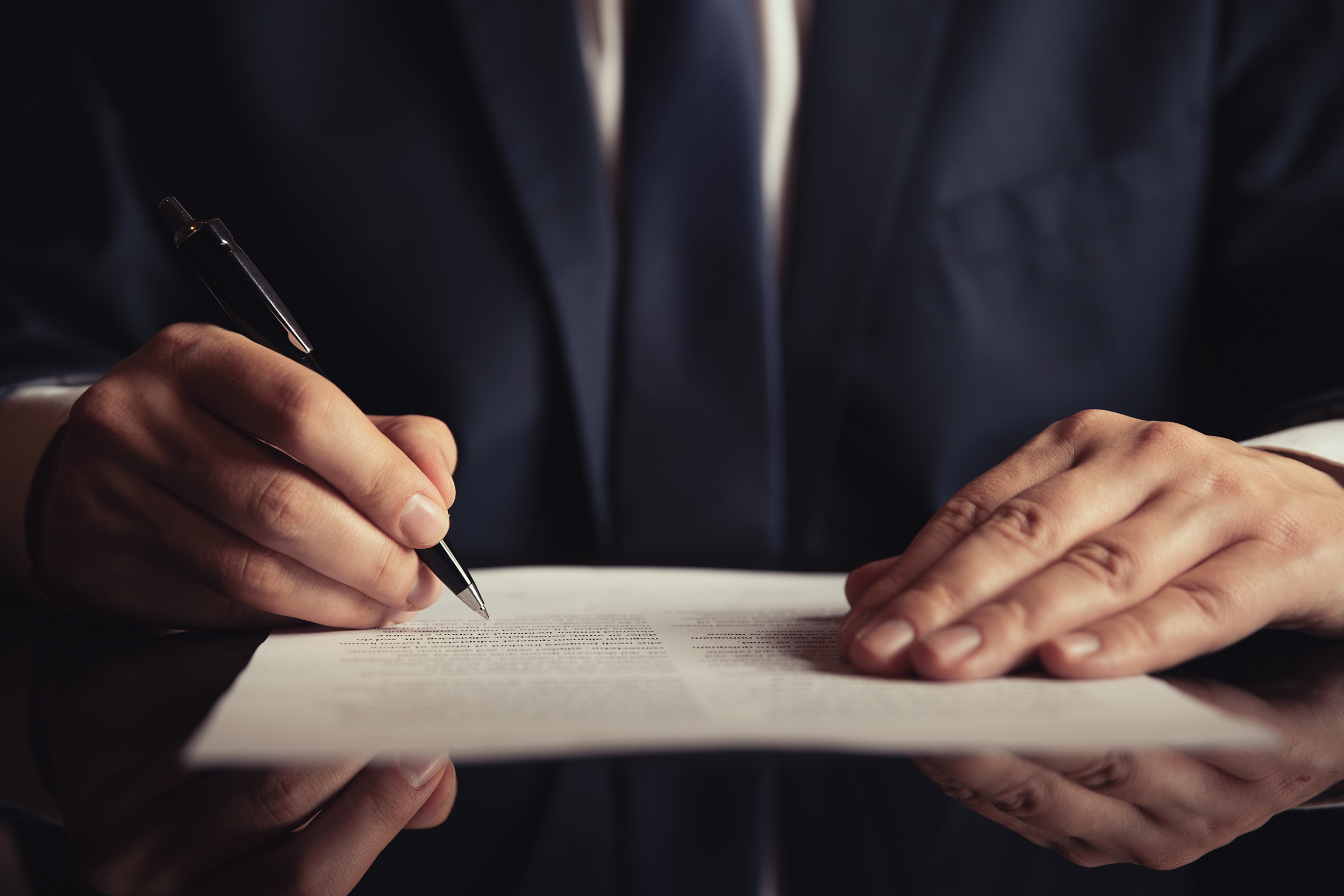 Person in a dark suit signing a document with a black pen, holding the paper steady with their other hand.