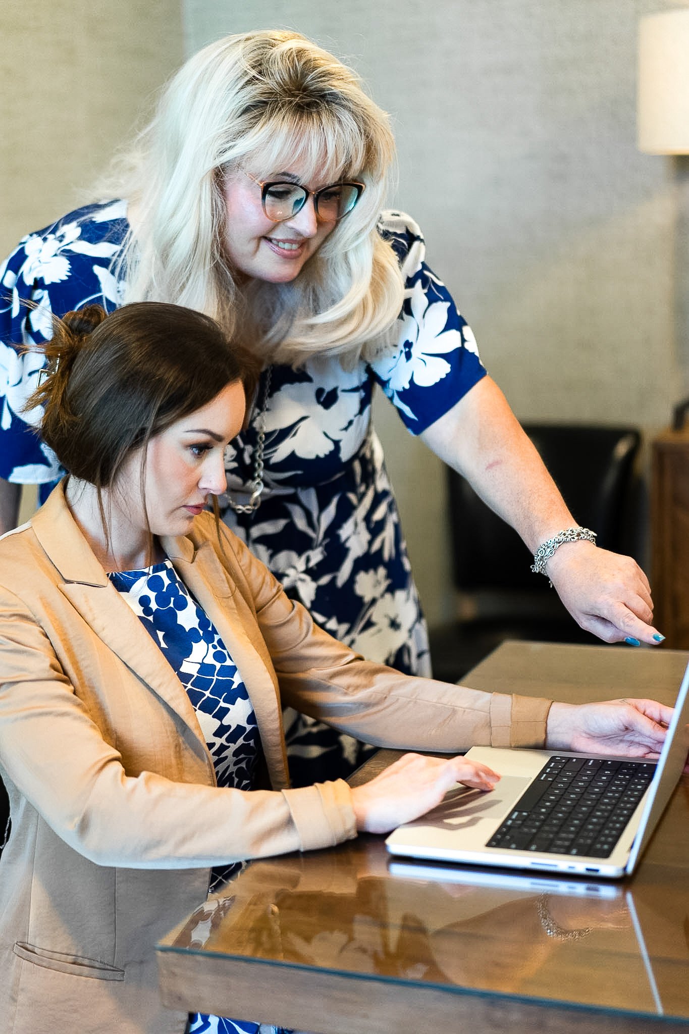 Two women, Brianna Peil and MJ Cooley, working together at a wooden table, one is sitting with a laptop and the other is standing and pointing at the screen.