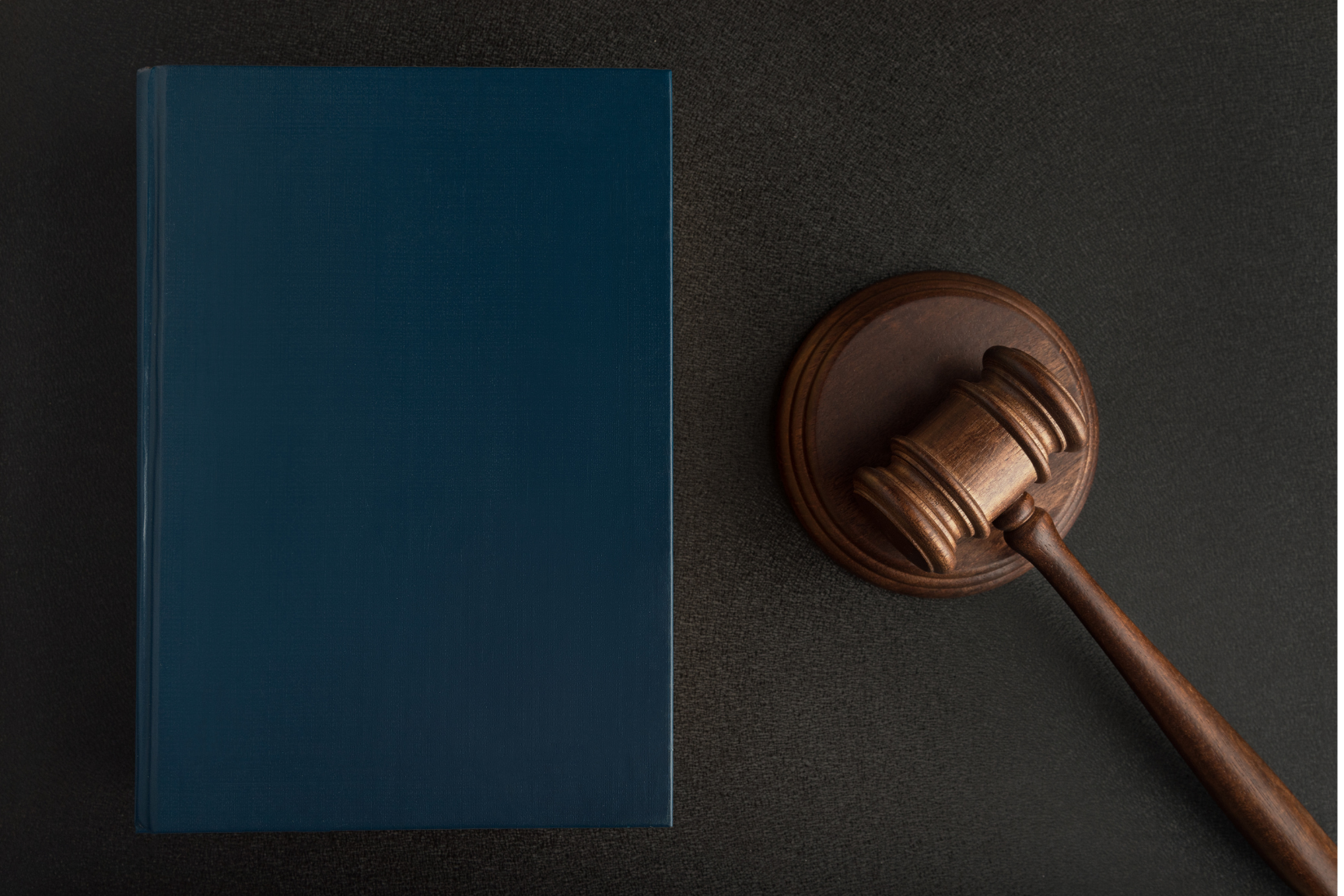 A wooden judge's gavel resting on a round sound block beside a closed blue hardcover book on a dark surface.