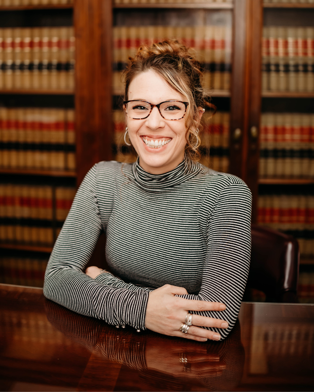 A woman, Jena Hutton, with glasses and earrings smiling, sitting at a polished wooden table with her arms crossed, in a room with bookshelves filled with legal books in the background.