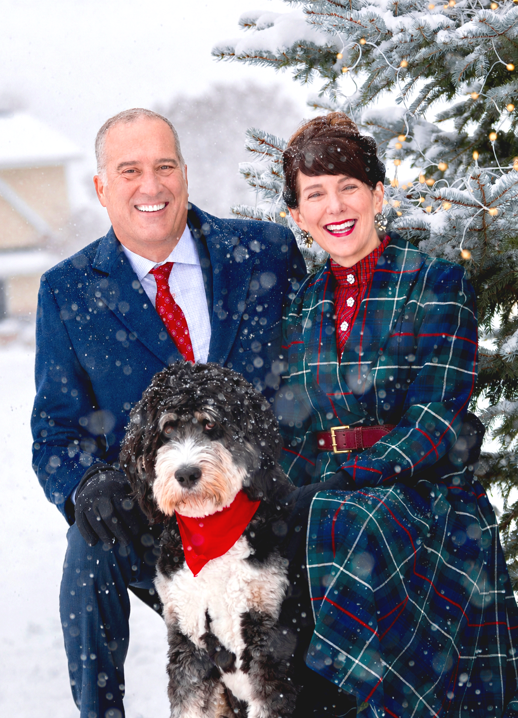 Carol and Brian Bruess in winter clothing posing outdoors with George, snow on a Christmas tree, and snow falling.