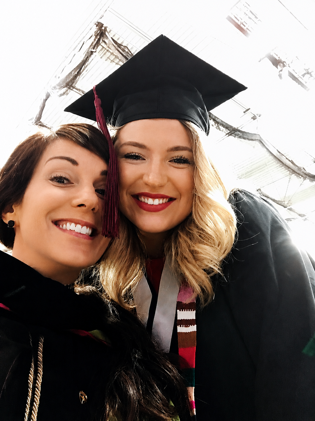 Carol Bruess celebrating graduation, with one wearing a cap and gown and the other dressed formally, smiling at the camera.