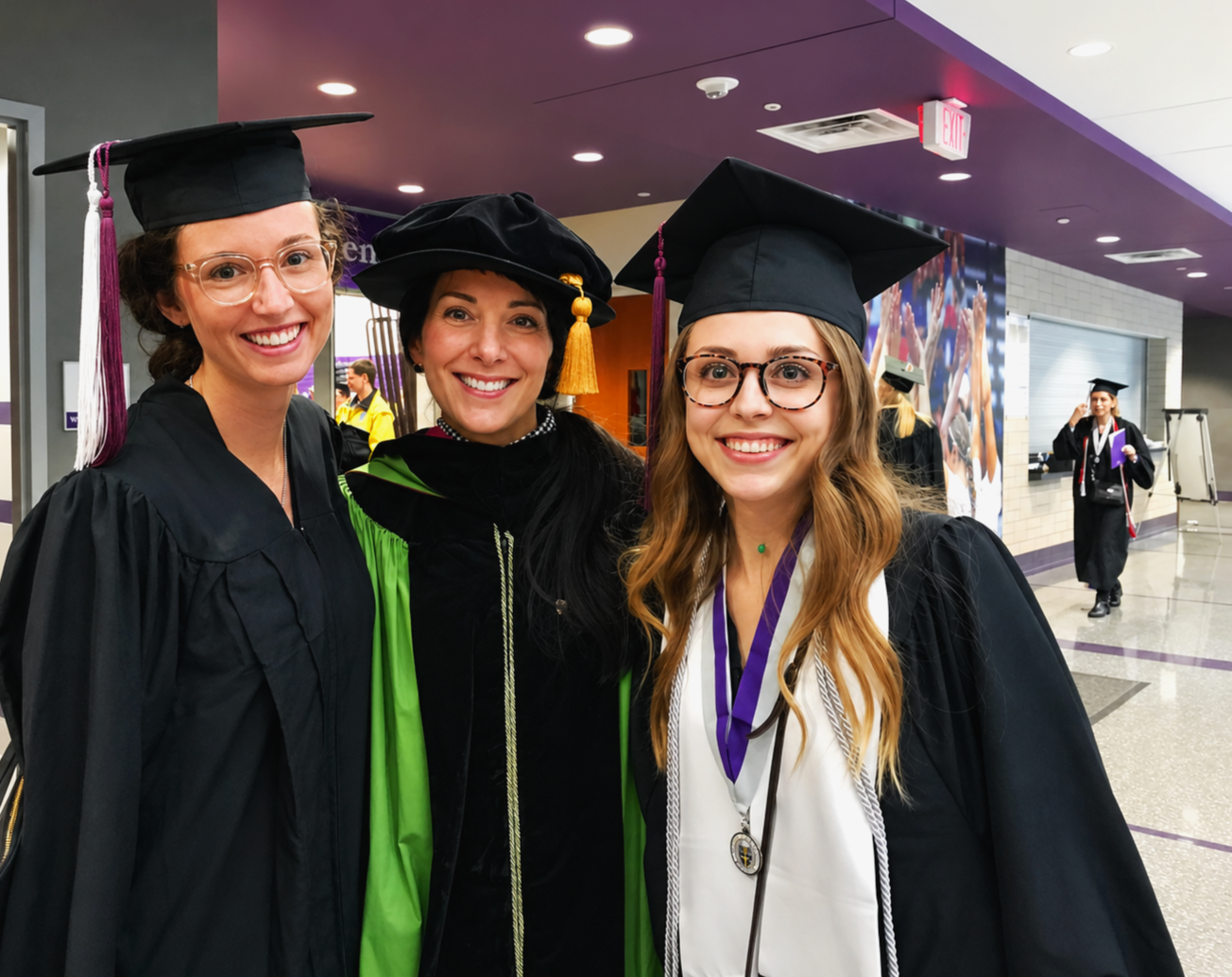 Carol Bruess and two other women in graduation caps and gowns smiling inside a building, with other graduates in the background.