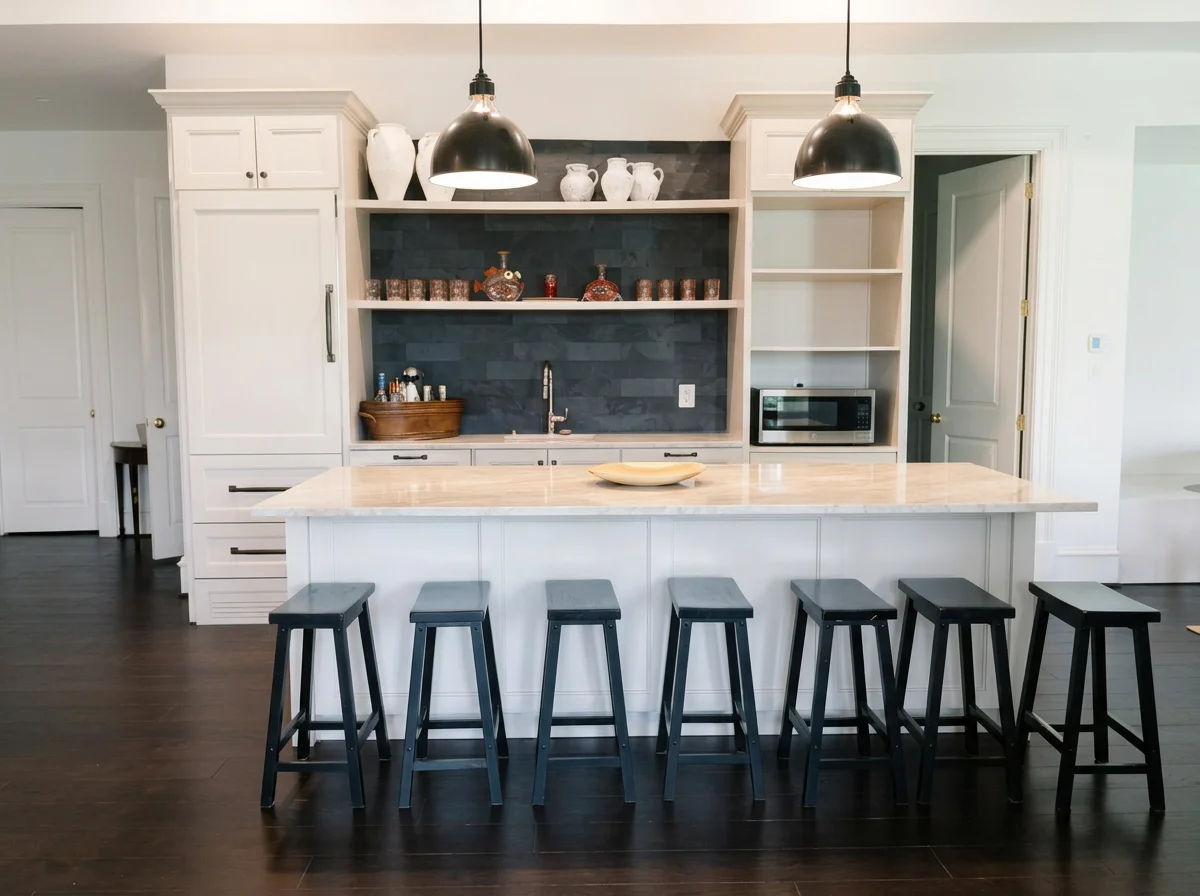 Modern kitchen with white cabinets, black bar stools, a large island with a light countertop, and a black accent wall with open shelves holding white vases, decorative jars, and small bottles. There are two black pendant lights hanging above the island, a microwave, and a door to the right.