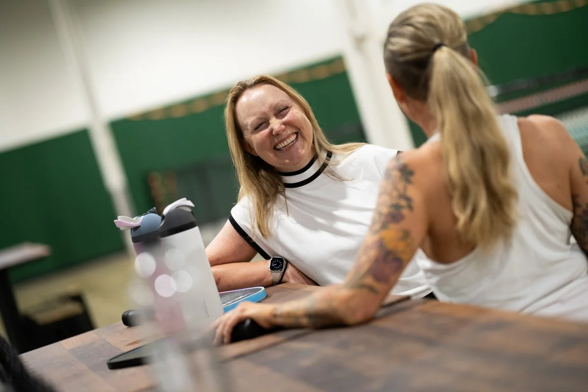 Two women talking and laughing at a table in an indoor setting, with a water bottle and a tablet visible on the table.