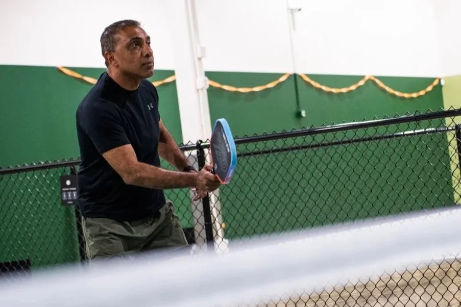 A middle-aged man with gray hair playing pickleball indoors, holding a paddle near the net, wearing a black t-shirt and beige shorts, with a green wall and yellow decorations in the background.