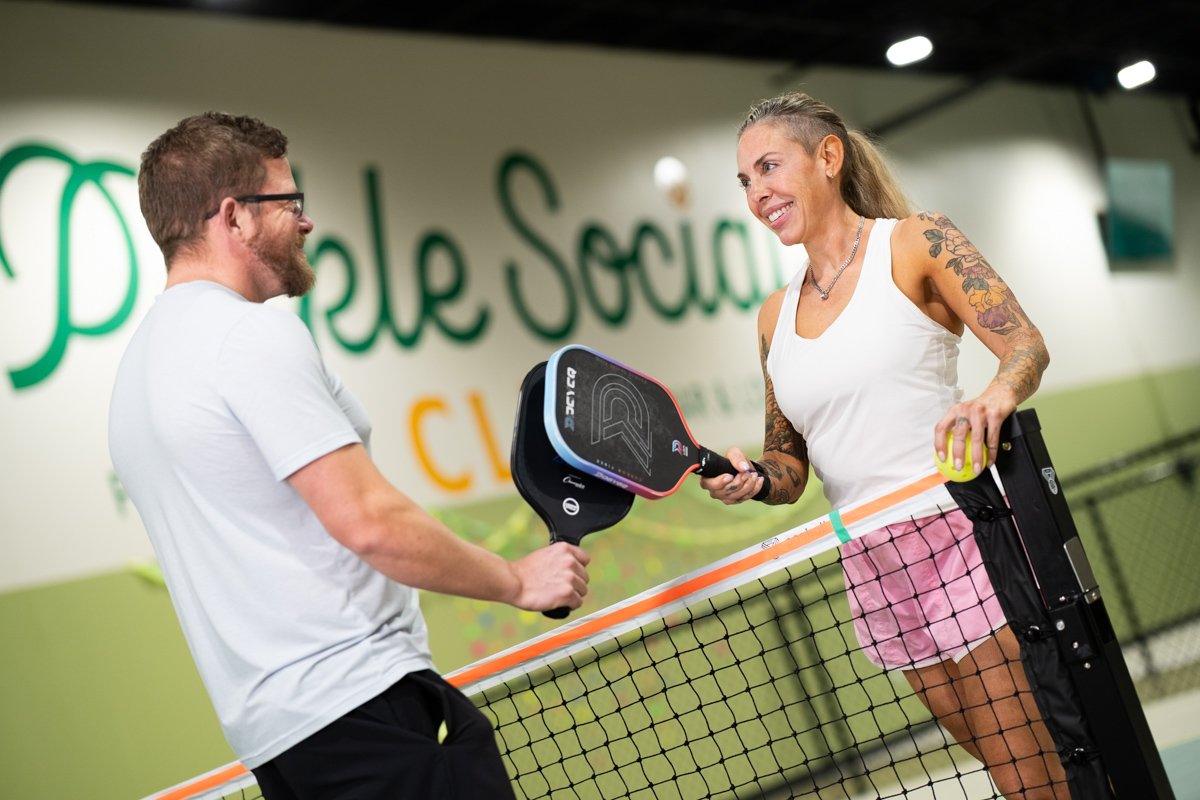 A woman with tattoos on her arms holding a pickleball paddle and an electric green pickleball while talking to a man with glasses and a beard holding another paddle, on a pickleball court at People's Social Club.