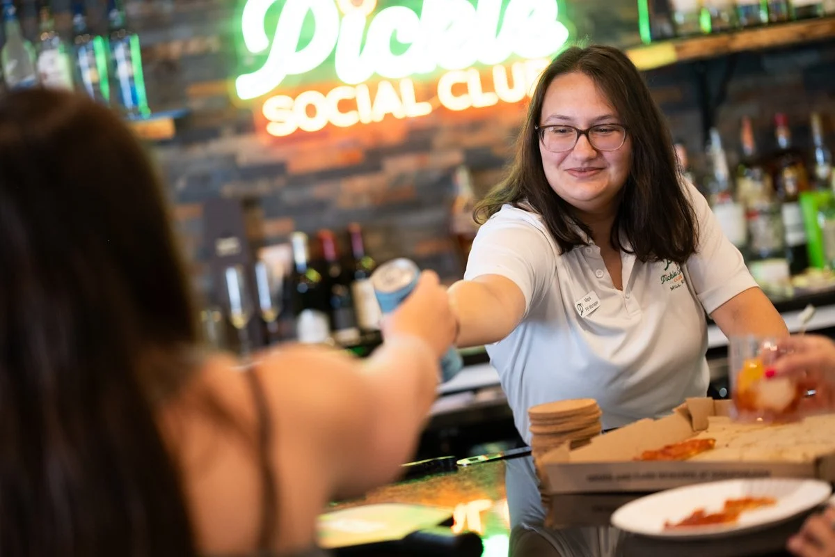 Barista handing a drink to a customer at a social club, with a pizza box and drinks on the counter, and a neon sign that reads 'Pizza Social Club' in the background.