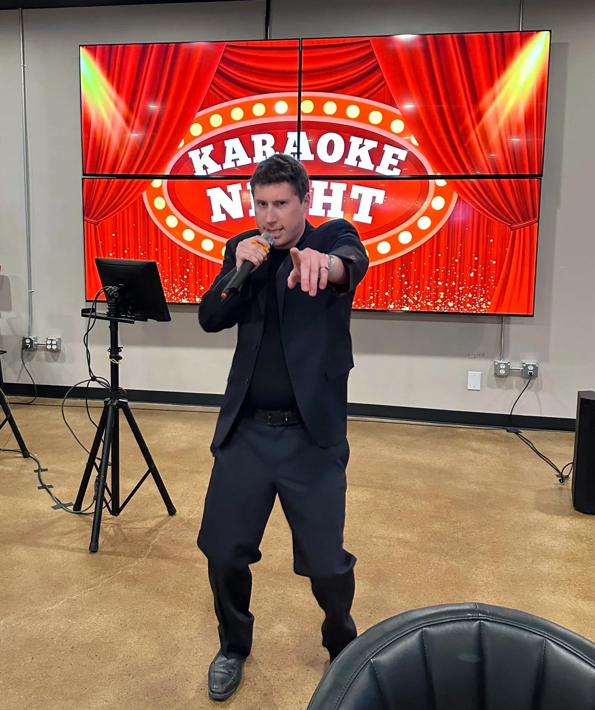 A man in black suit pointing at the camera while holding a microphone with his right hand in front of a red curtains backdrop with the words 'Karaoke Night' and stage lights displayed on a large screen behind him.