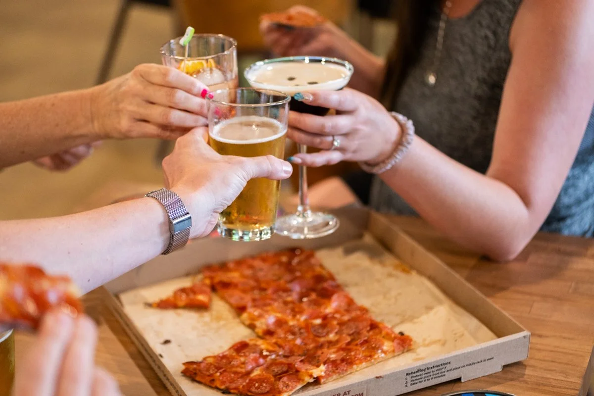 Group of people raising glasses of beer and cocktails for a toast around a table with a pizza on a tray.