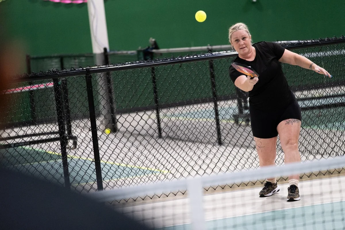 Woman in black athletic clothes playing pickleball on an indoor court, preparing to hit a yellow ball.