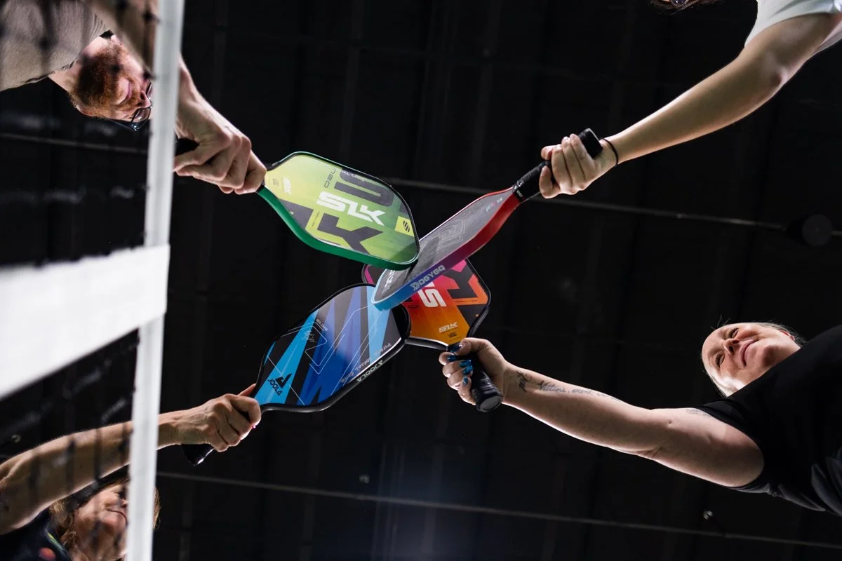 Four people clinking colorful pickleball paddles together in an indoor setting.