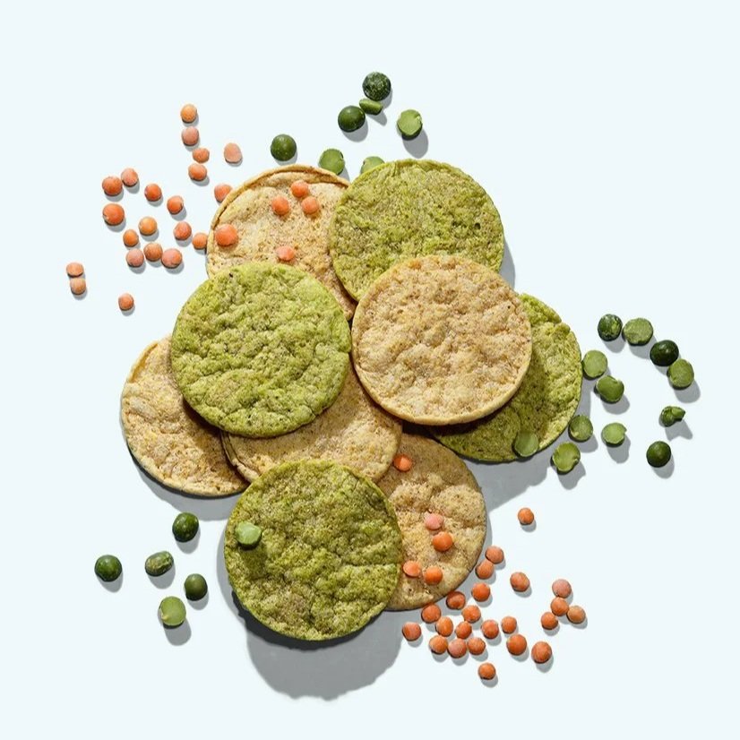 A pile of green and beige crunchy crisps with scattered red lentils and green peas on a light blue background