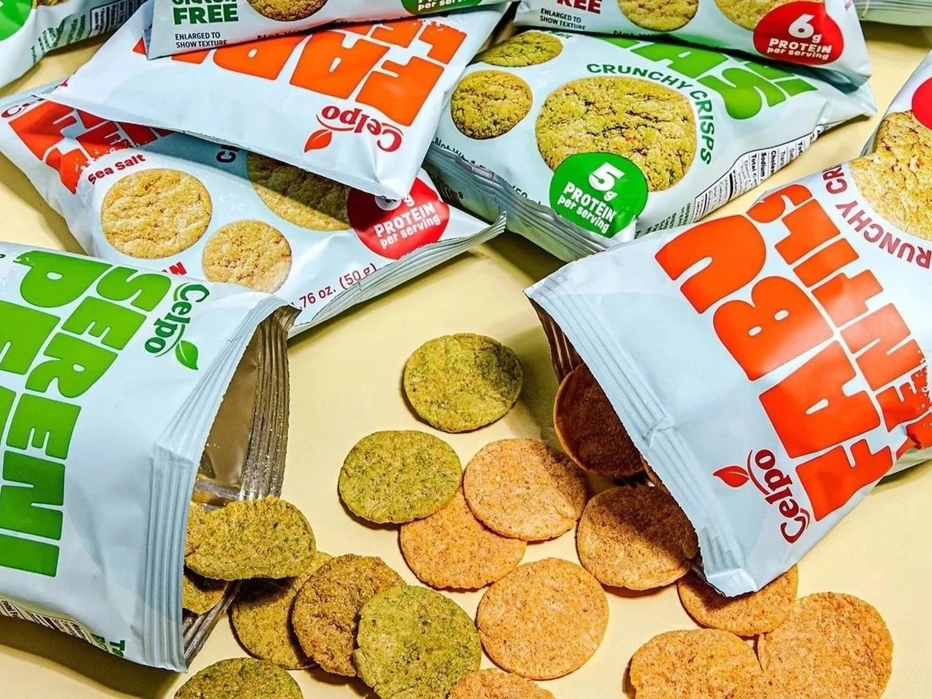 Multiple bags of Celpo FabuLentils™ and SereniPeas™ Crunchy Crisps in Sea Salt flavor, with some chips spilled onto a light-colored surface.