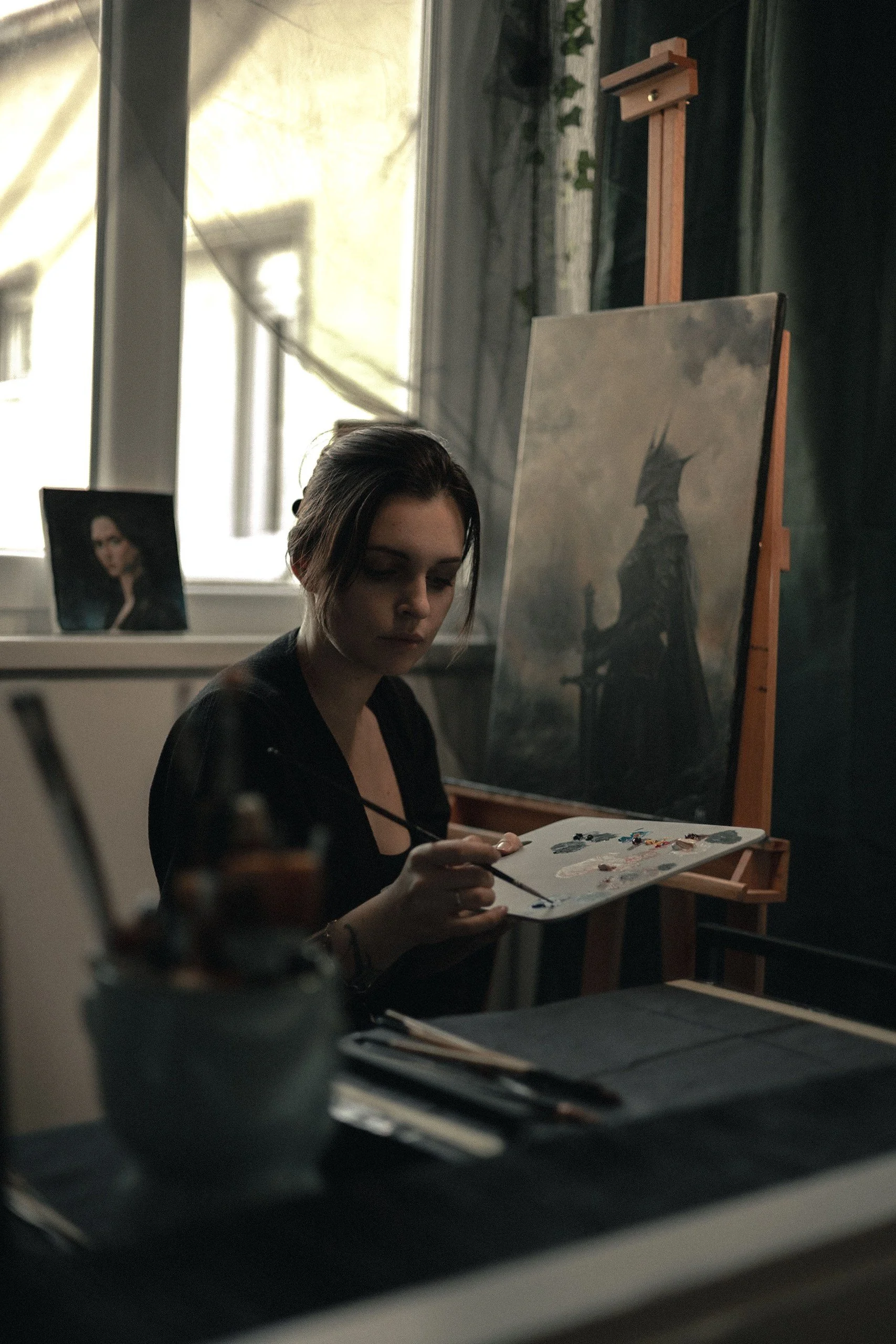 A woman with short hair drying paint on a canvas in a well-lit art studio, with an easel and art supplies nearby.