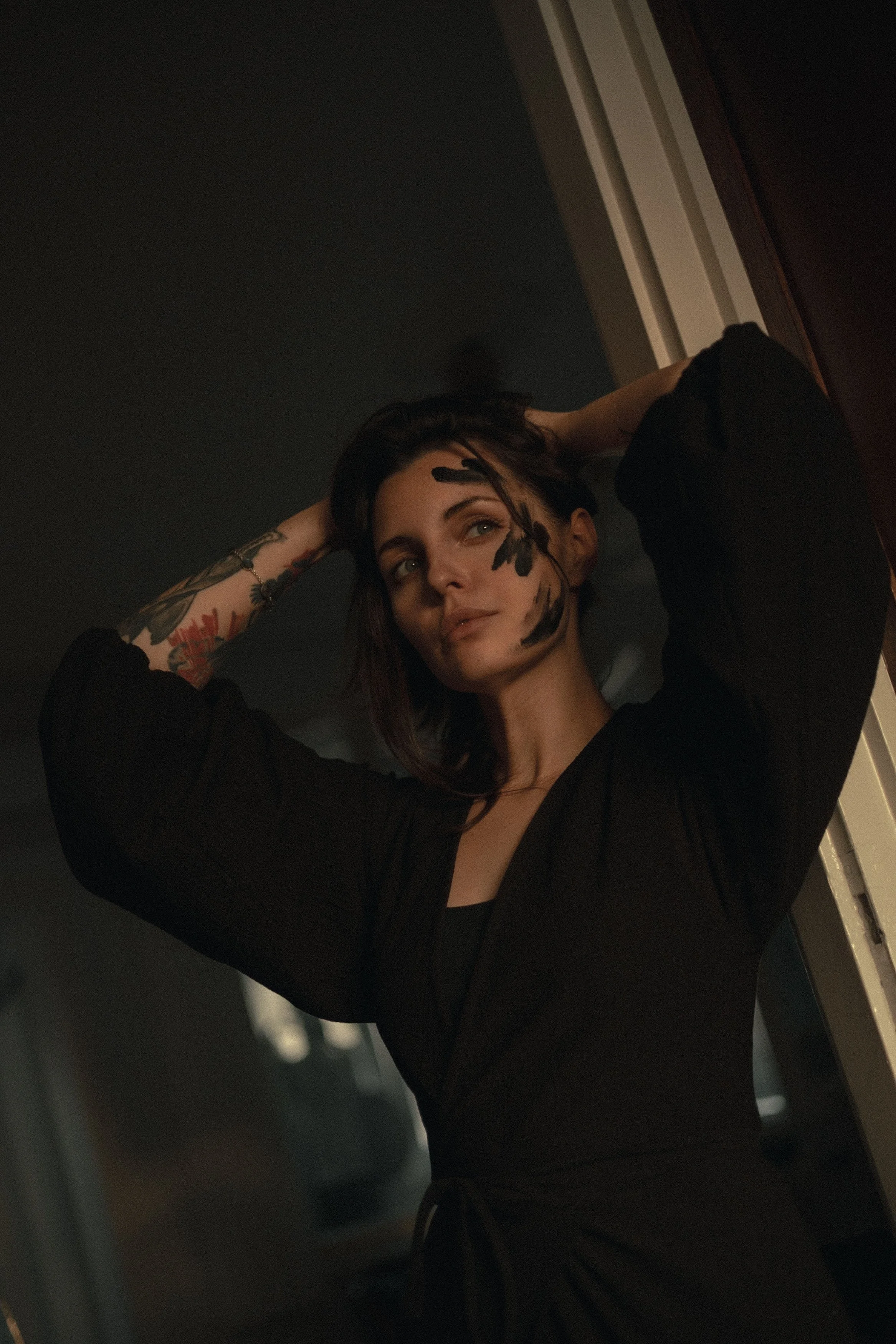 A woman with shoulder-length dark hair and tattoos on her left arm, wearing a black top, standing in a dimly lit indoor setting, looking slightly upward with her hands positioned behind her head.