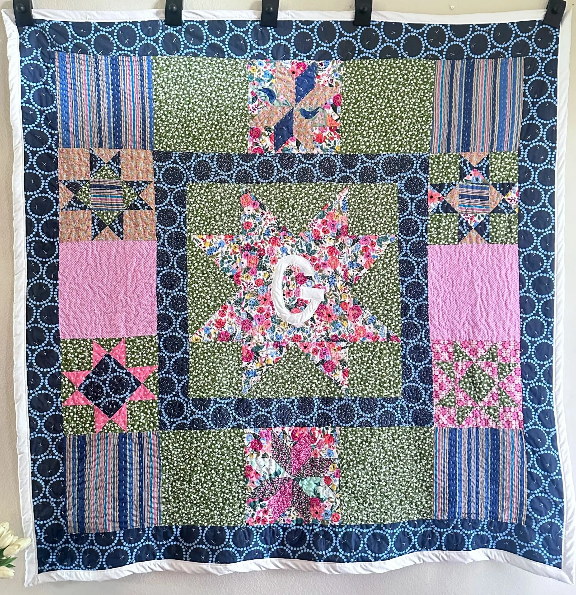 A quilt hanging on a wall with a black, blue, pink, and floral pattern.