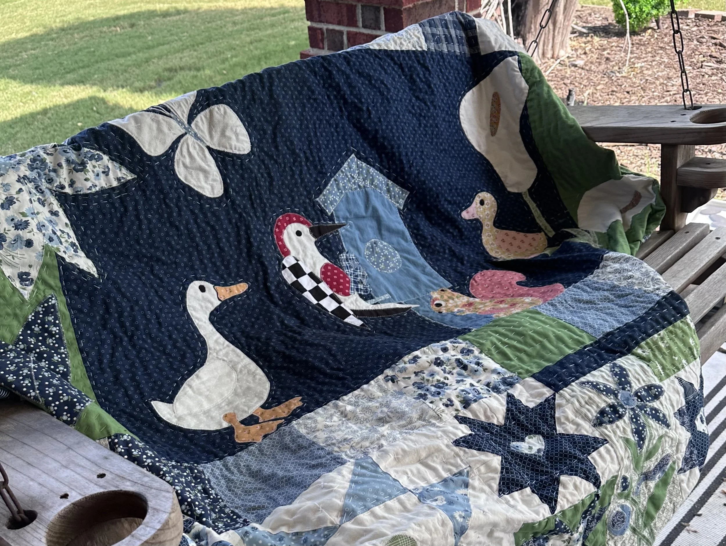 A colorful quilt with a design featuring a duck, duckling, and squirrel is draped over a wooden porch swing.