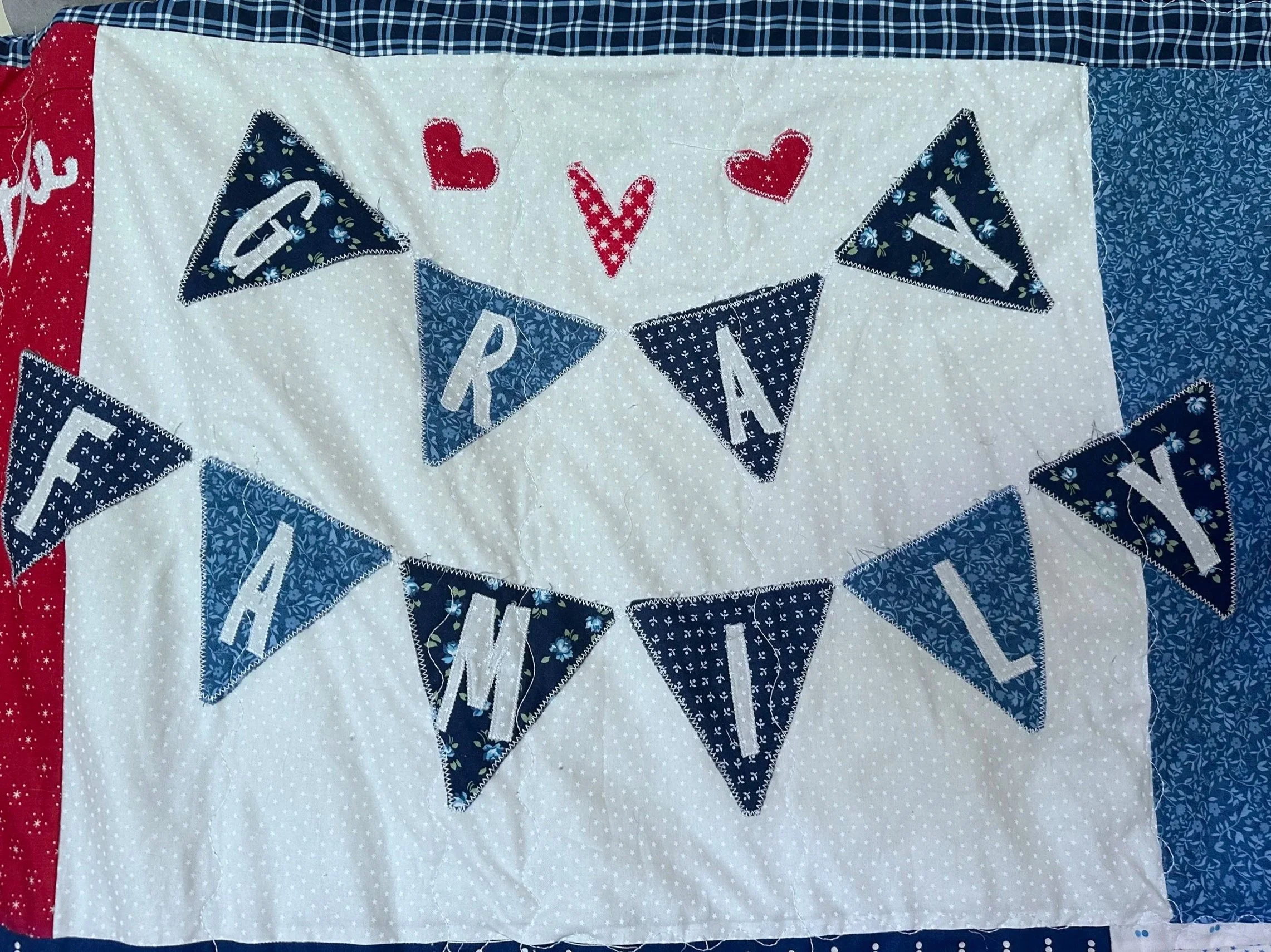 A quilted fabric banner with white background, decorated with blue and red patterned bunting flags spelling out 'Gray Family' and small red hearts.