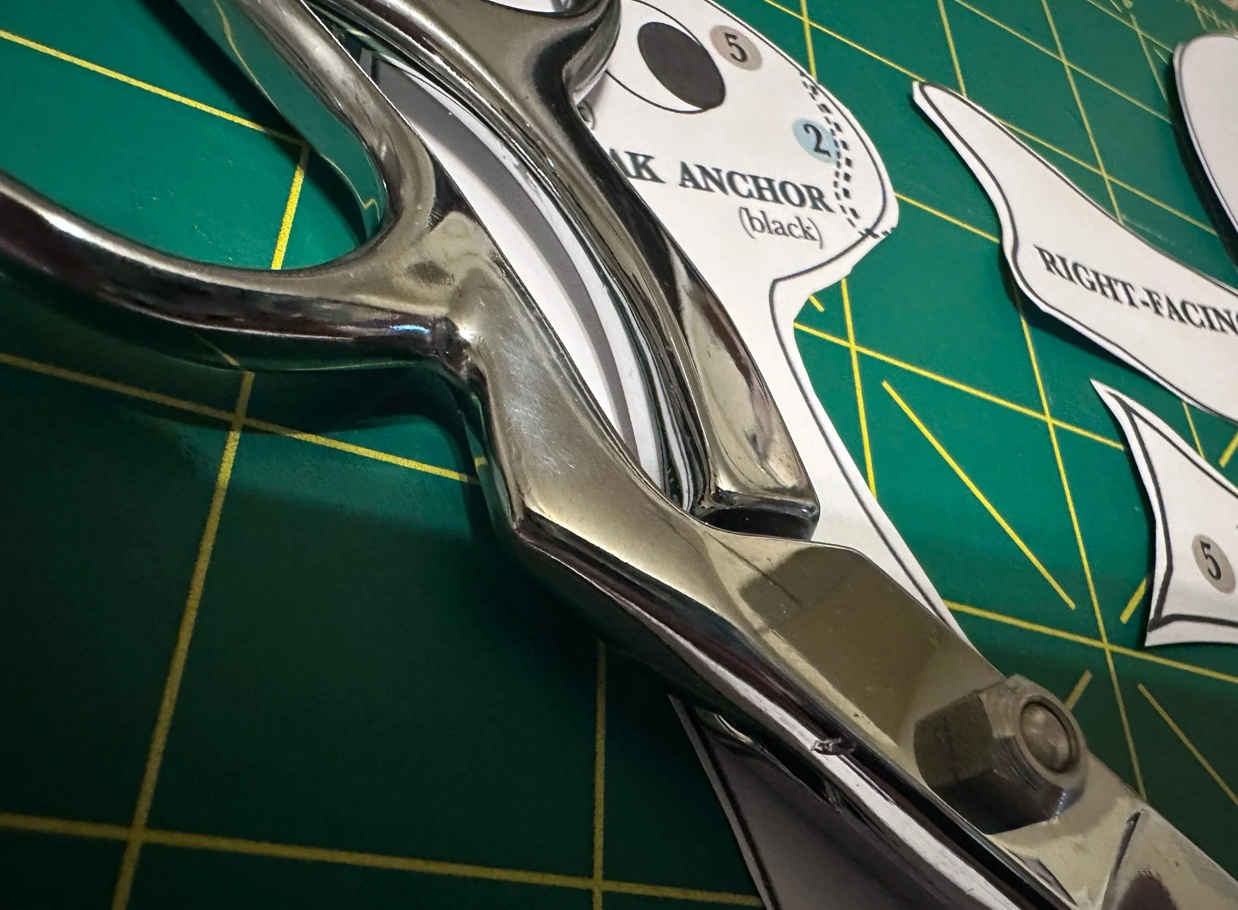 Close-up of a shiny metallic golf club head on a green cutting mat with yellow grid lines. Attached paper labels or templates read 'RIGHT-HEEL' and '1K ANCHOR (black)'.