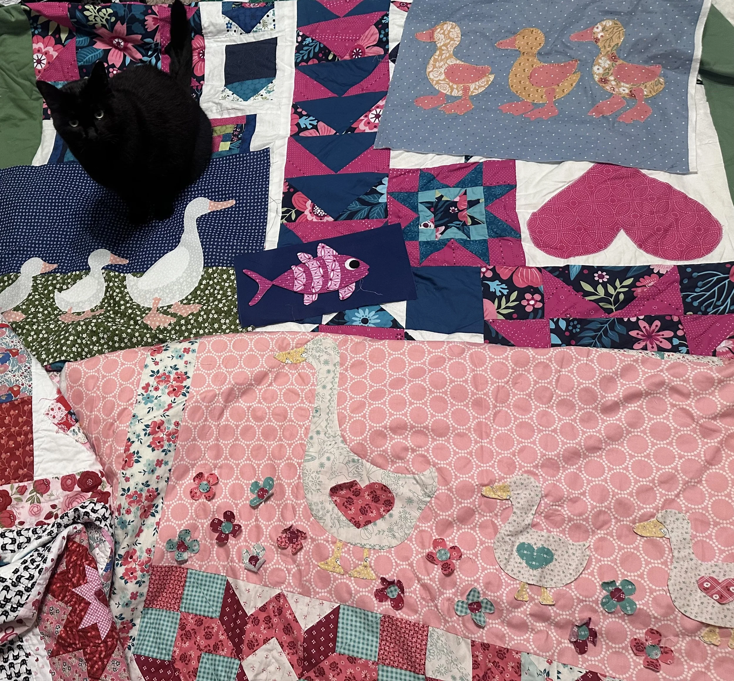 Colorful patchwork quilt with various patterns and designs, including swans, ducks, fish, hearts, and floral patterns, along with a black cat sitting on the quilt.