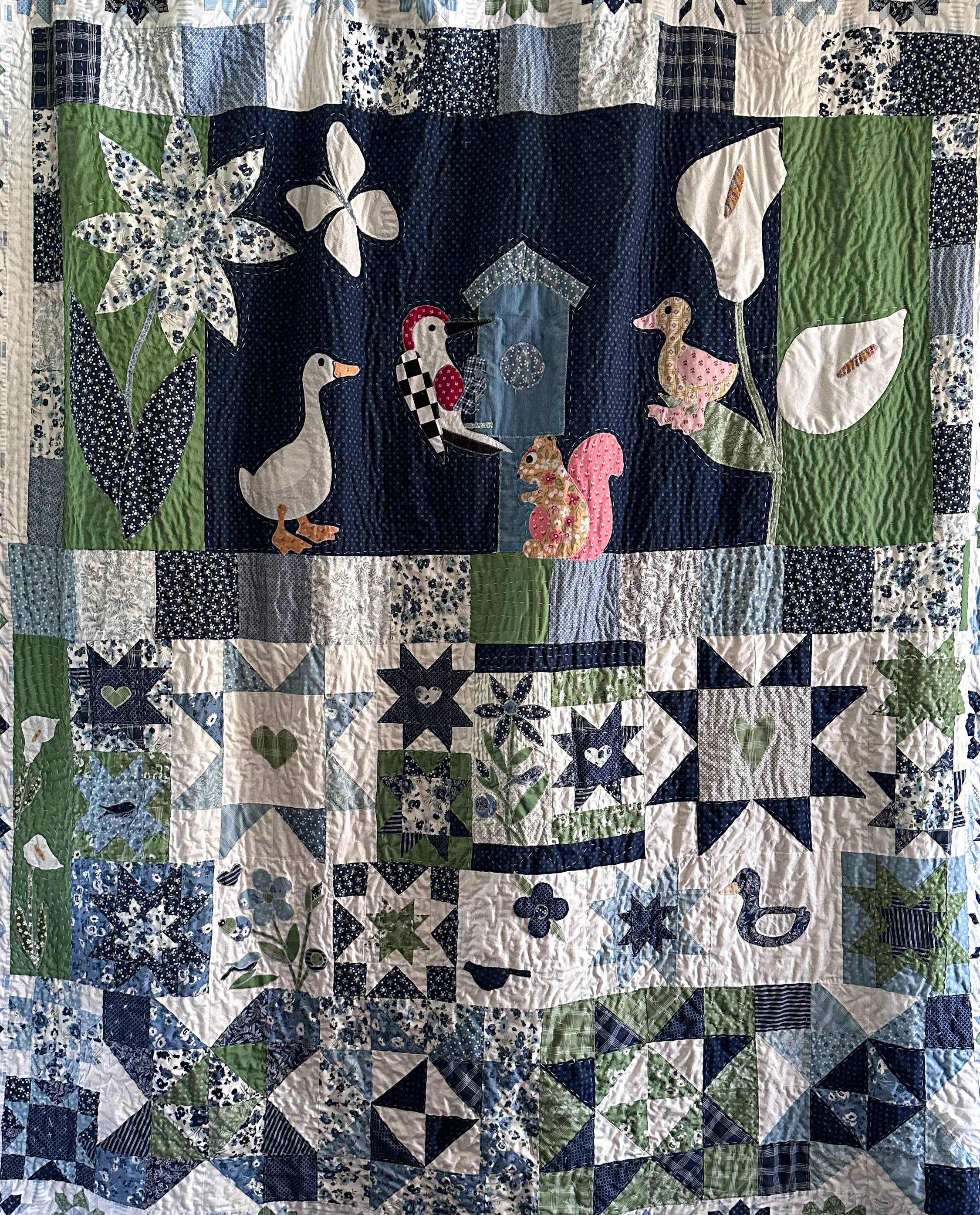 Close-up of a quilt featuring a dark blue background with whimsical animal and nature appliqué, including ducks, a squirrel, a duckling, butterflies, flowers, leaves, and a birdhouse, with a border of various blue and green patterned fabric squares.