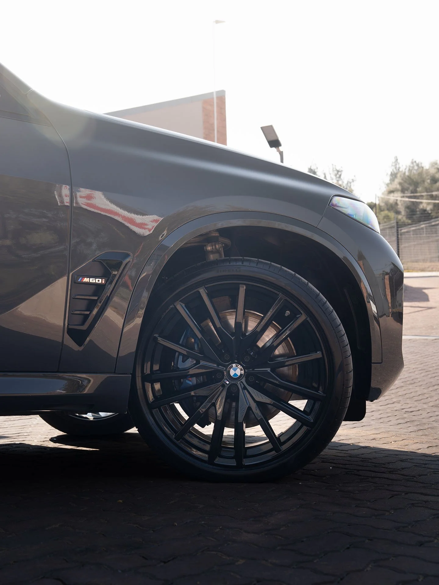 Close-up of the front part of a black BMW M60i car, showing the wheel, tire, and part of the front fender with the BMW logo and M60i badge, parked on a paved area.