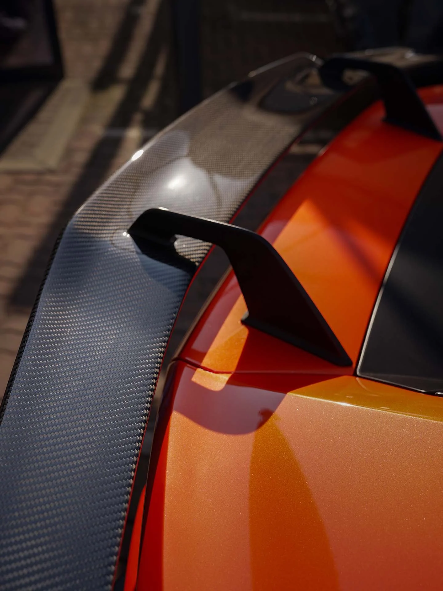 Close-up of the rear wing of a sports car, showing a carbon fiber diffuser with a black fin and an orange body.