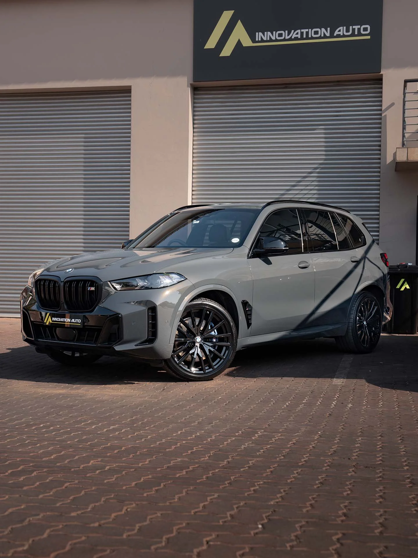 A silver BMW X5 M parked in front of a building with a sign that reads ``Innovation Auto'' and a rolling metal door. The car has black rims and tinted windows.
