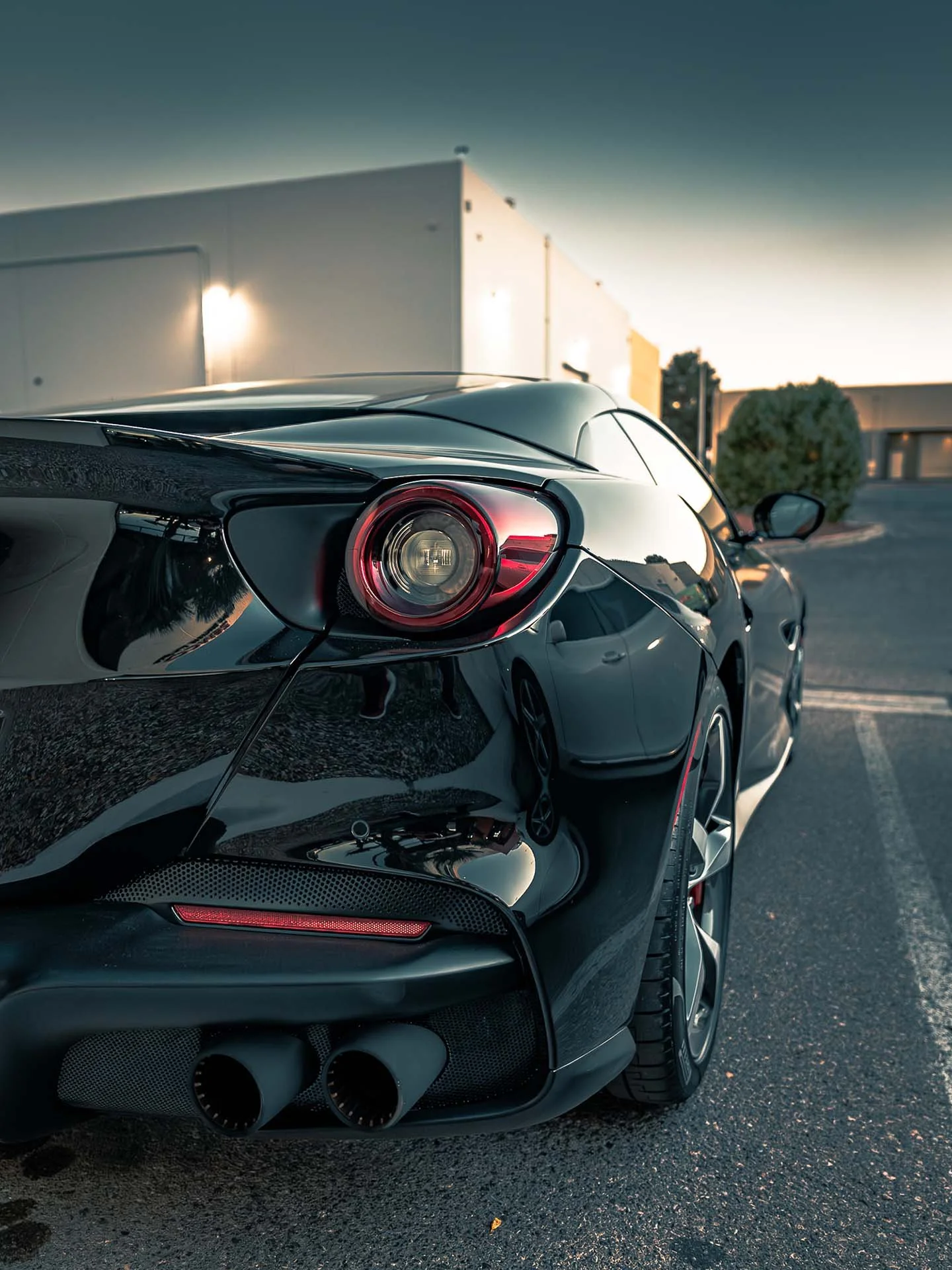 Close-up of the rear of a black sports car, showing sleek tail lights, a large exhaust pipe, and reflections of the surrounding area, with a building and trees in the background during sunset.