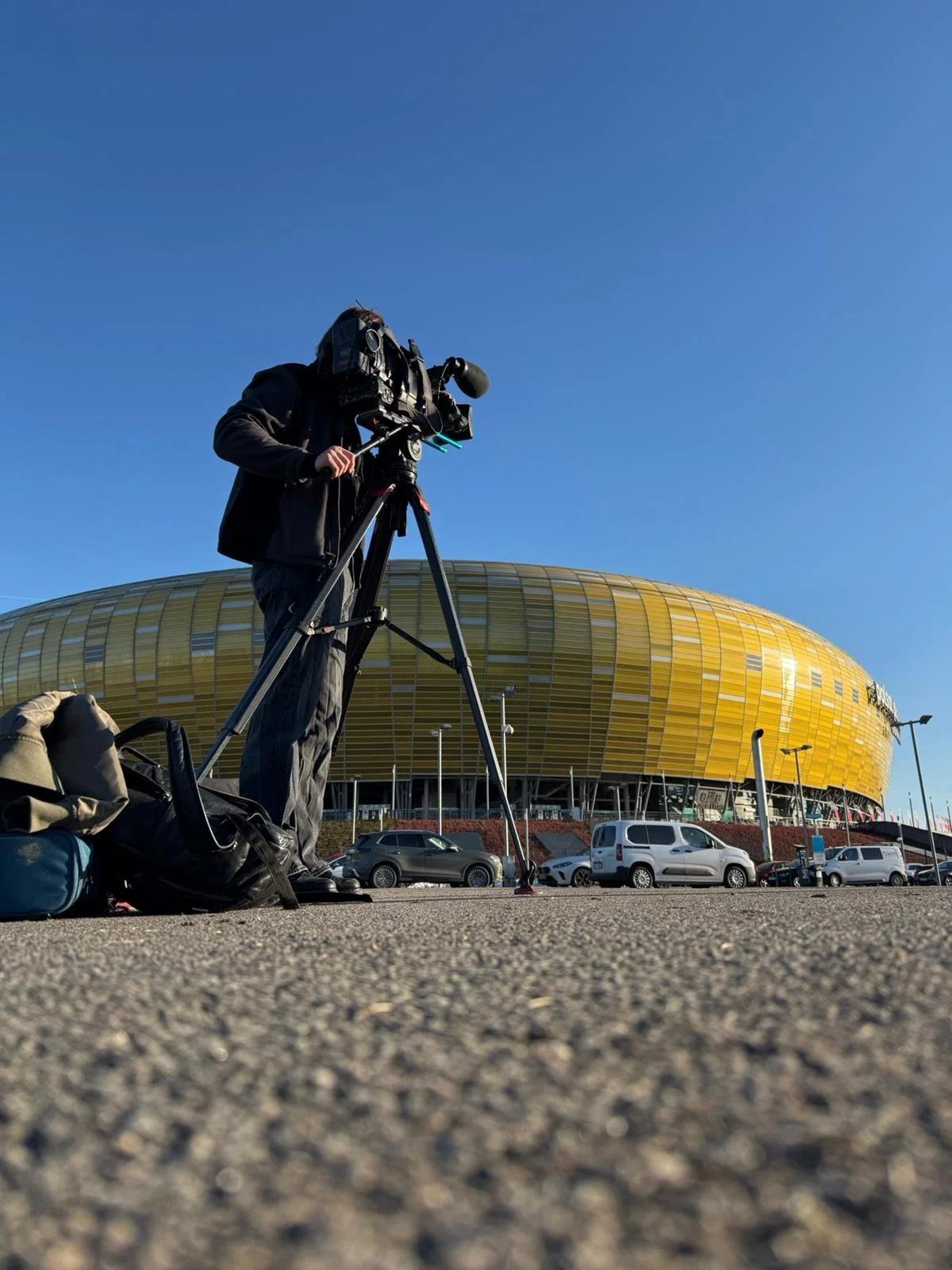 A person operating a professional video camera on a tripod outside in front of a large yellow building with a rounded design, under a clear blue sky.