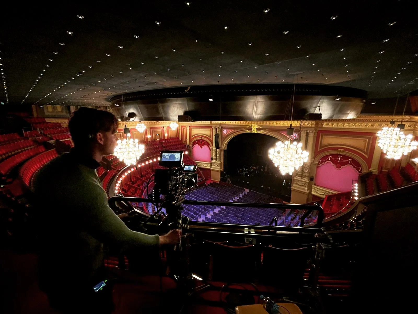A person operating camera equipment in a grand, empty theater or opera house with red velvet seats, ornate gold decorations, and large chandeliers hanging from the ceiling.