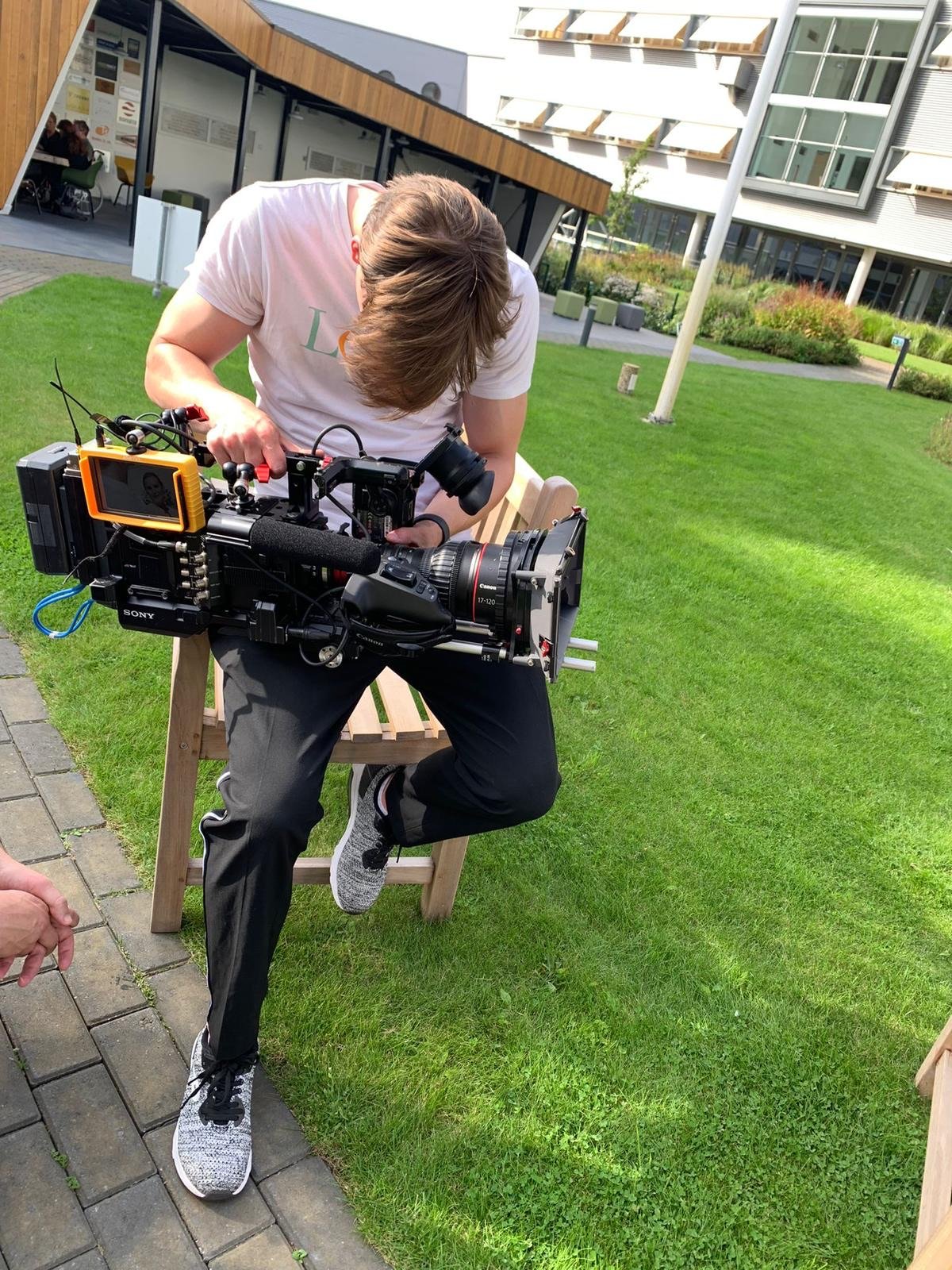 A person adjusting a professional video camera outdoors on a grassy area with modern buildings in the background.