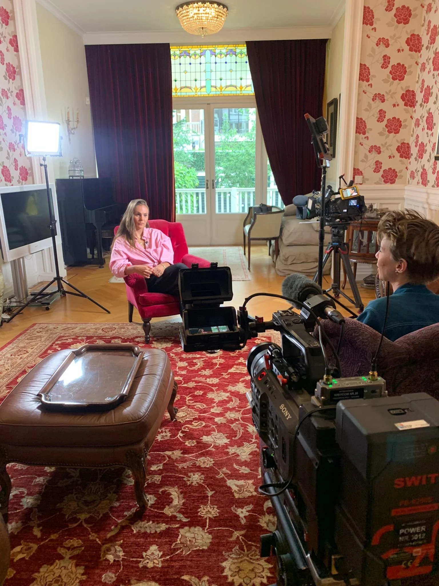 Scene of a filming set in a living room with a woman sitting on a red armchair, surrounded by professional video equipment, cameras, lighting, and crew members.